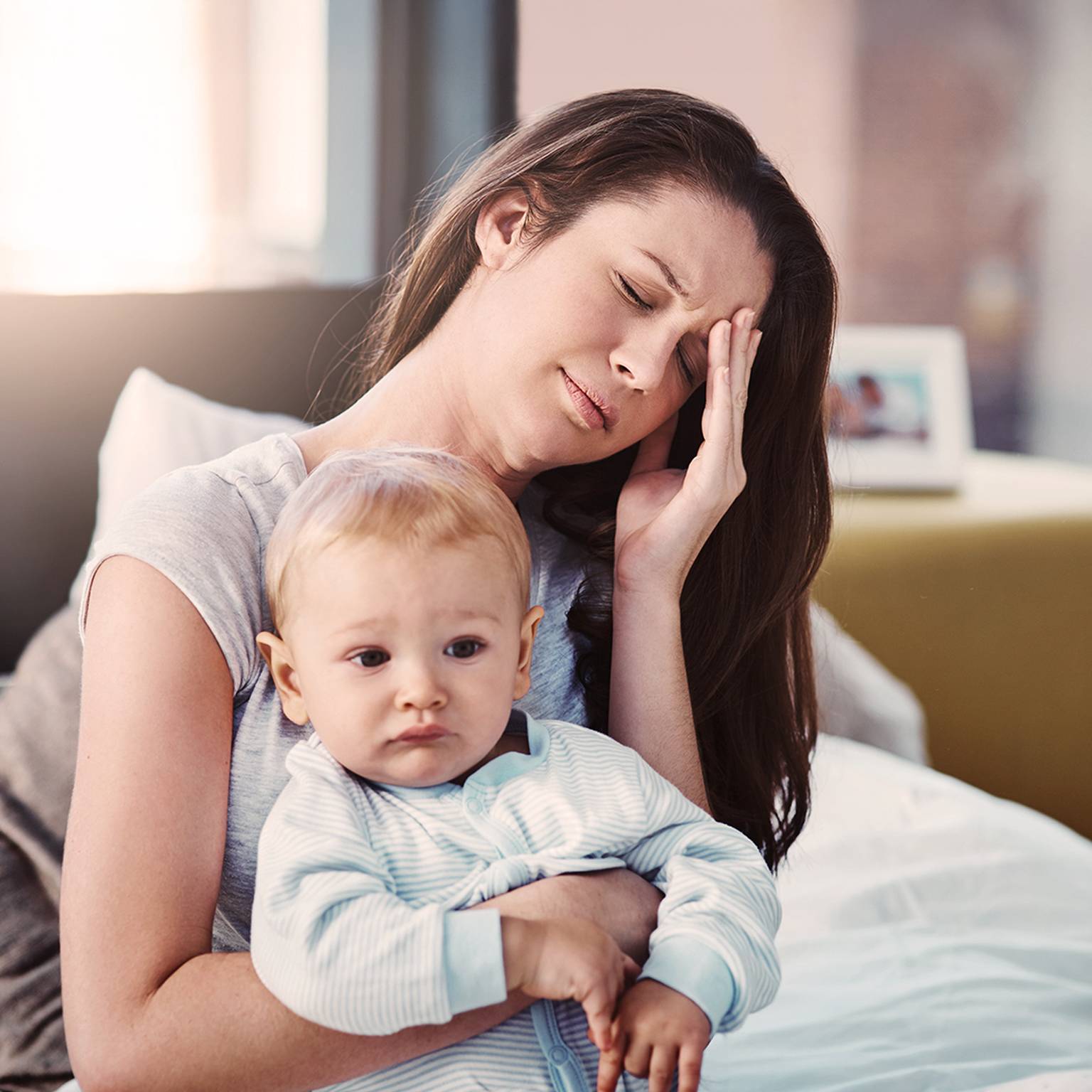 Eine müde Frau, die auf einem Bett sitzt, ein Baby im Arm hält, die Hand auf der Stirn hat und gestresst aussieht.
