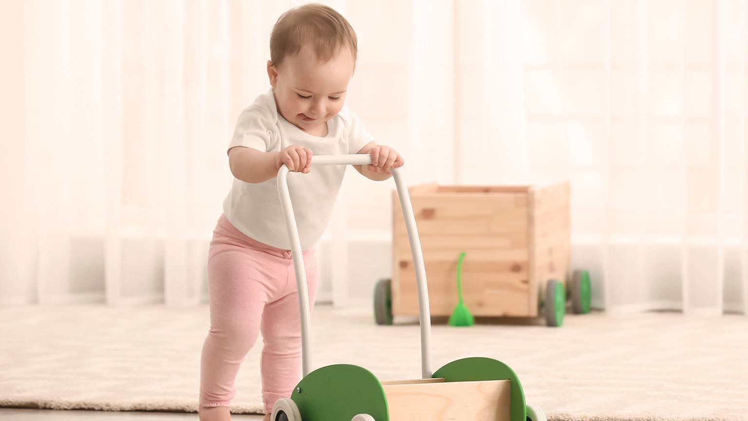 Ein Baby in rosafarbener Hose und weißem Hemd schiebt ein grünes Lauflernspielzeug aus Holz über einen mit Teppich ausgelegten Boden in einem hellen Raum.