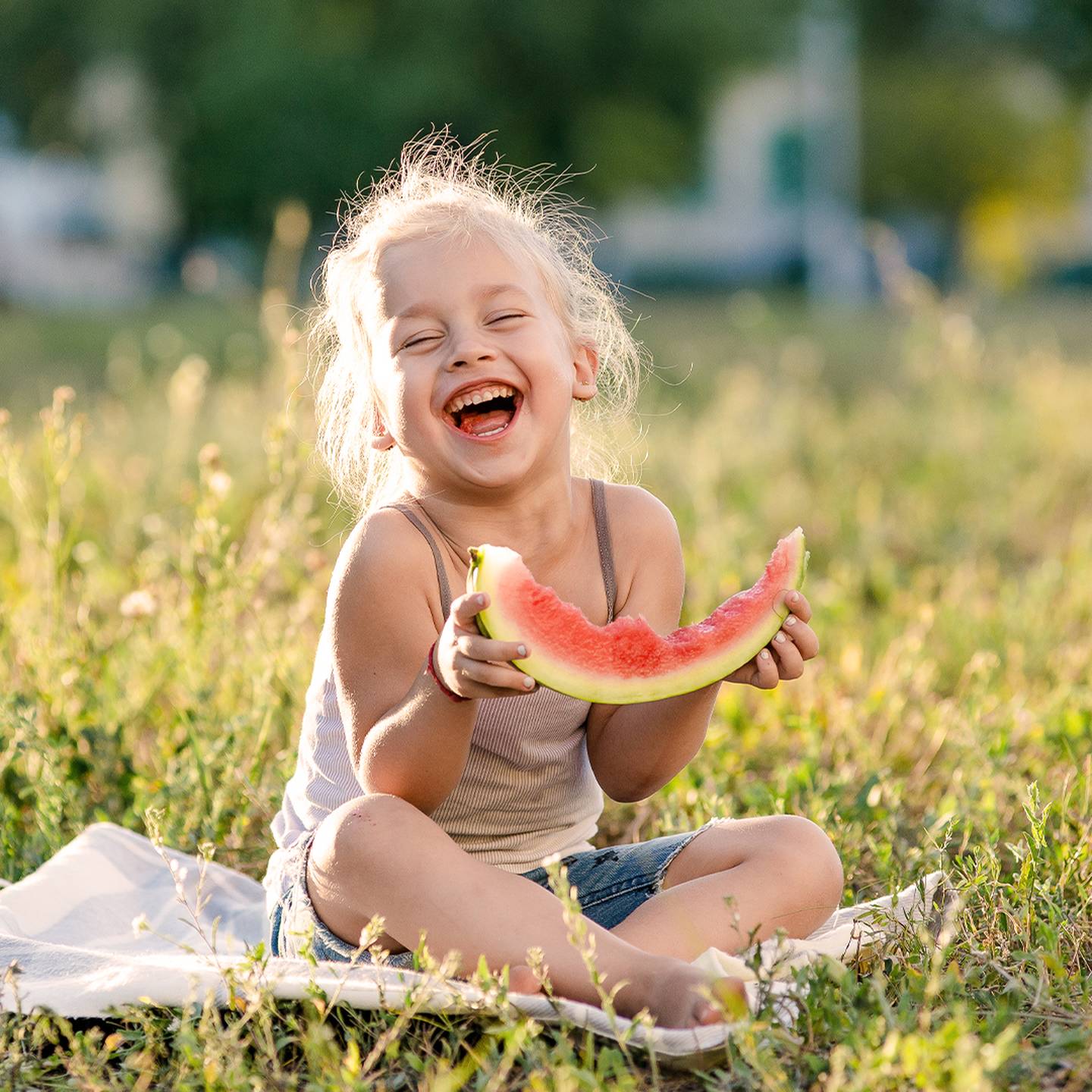 Ein fröhliches Kind sitzt lachend im Gras und hält ein Stück Wassermelone in der Hand. Sonnenlicht erhellt die Szene und schafft eine warme, fröhliche Atmosphäre.