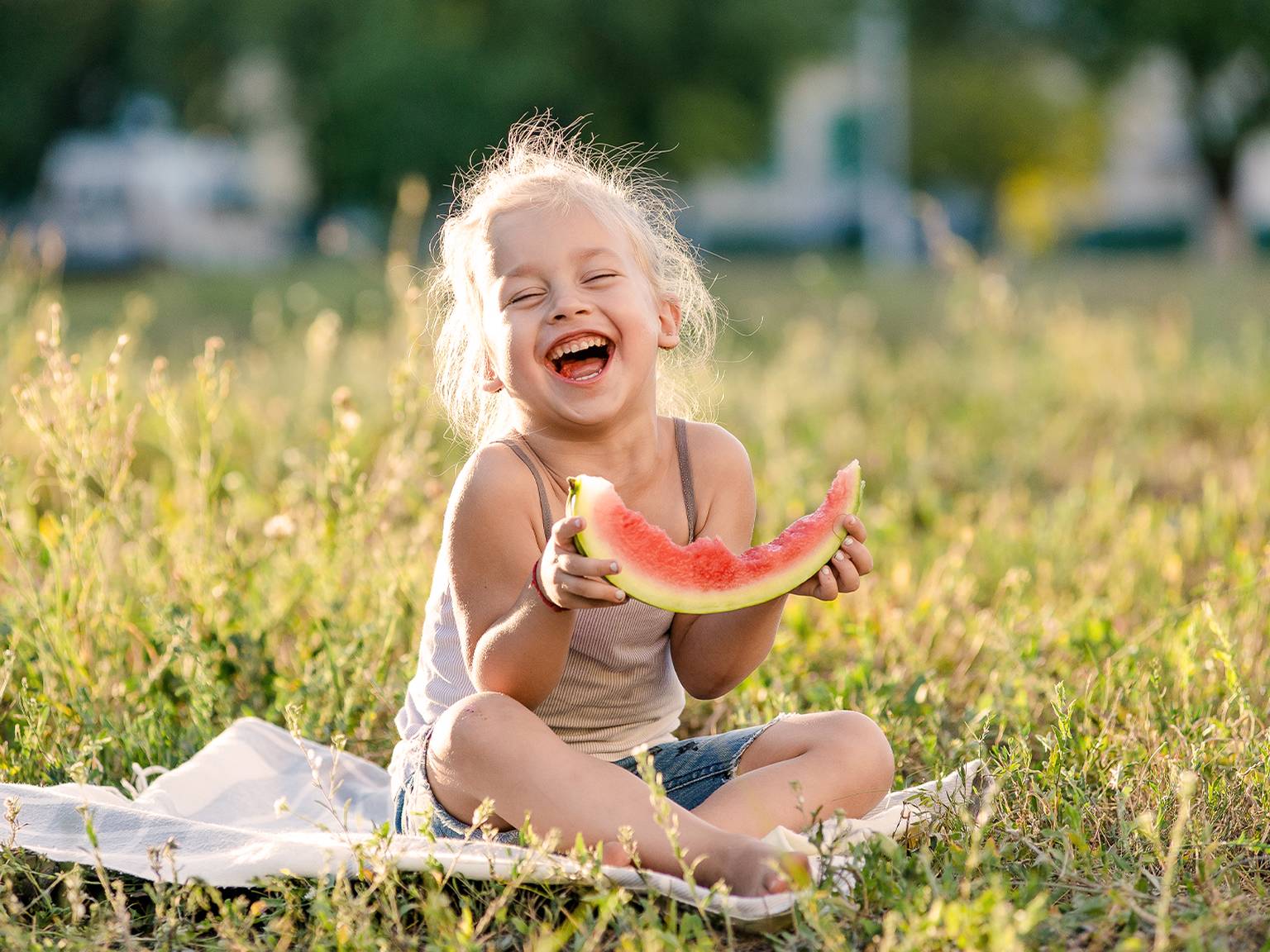 Ein fröhliches Kind sitzt lachend im Gras und hält ein Stück Wassermelone in der Hand. Sonnenlicht erhellt die Szene und schafft eine warme, fröhliche Atmosphäre.