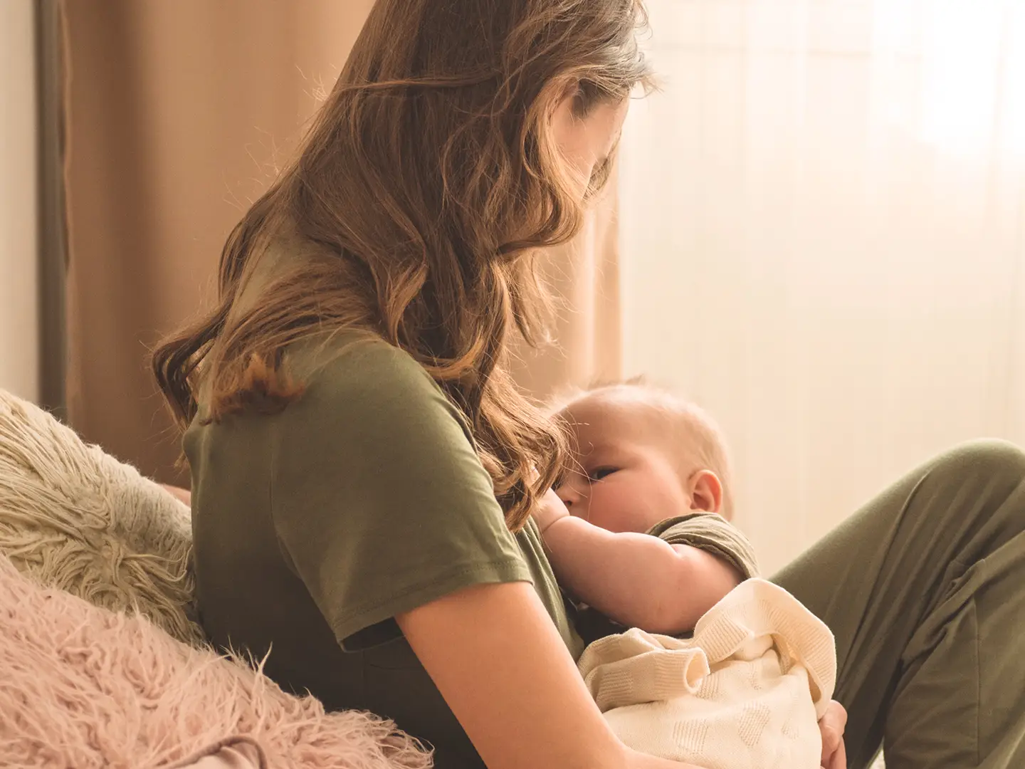 Eine Frau mit langen Haaren und einem grünen Hemd stillt ein in eine leichte Decke gewickeltes Baby und sitzt auf einem gemütlichen, sanft beleuchteten Sofa.