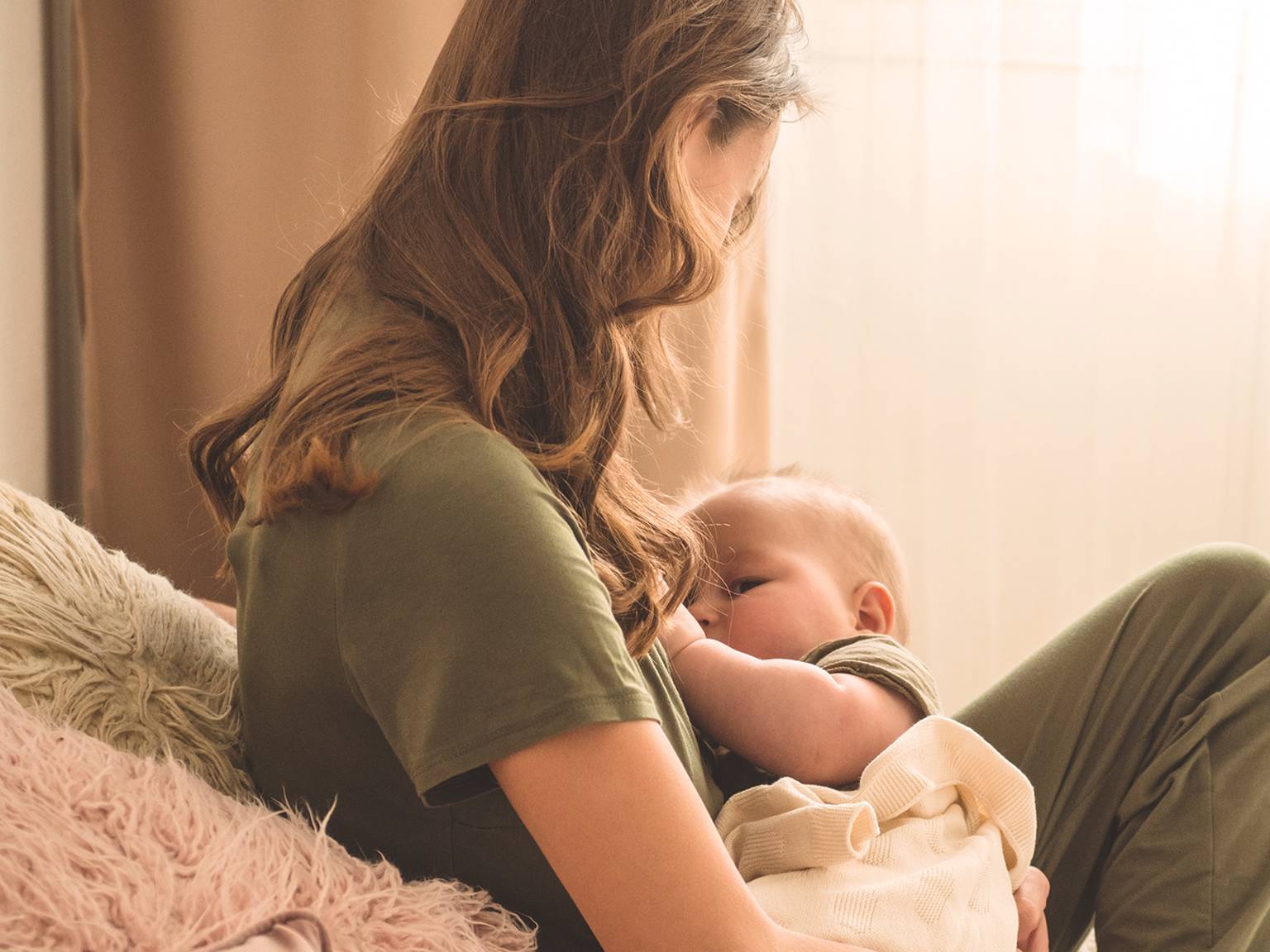 Eine Frau mit langen Haaren und einem grünen Hemd stillt ein in eine leichte Decke gewickeltes Baby und sitzt auf einem gemütlichen, sanft beleuchteten Sofa.