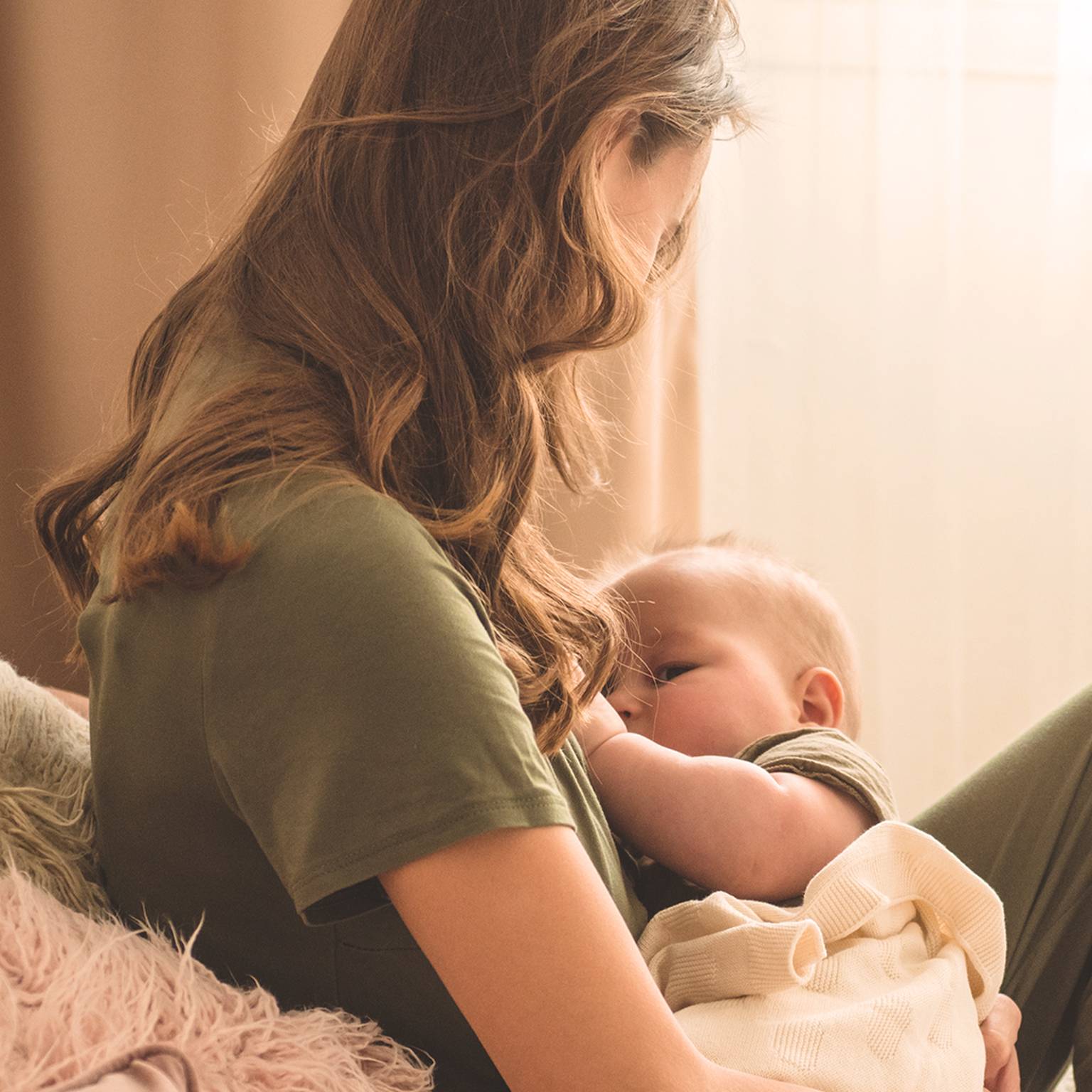 Eine Frau mit langen Haaren und einem grünen Hemd stillt ein in eine leichte Decke gewickeltes Baby und sitzt auf einem gemütlichen, sanft beleuchteten Sofa.