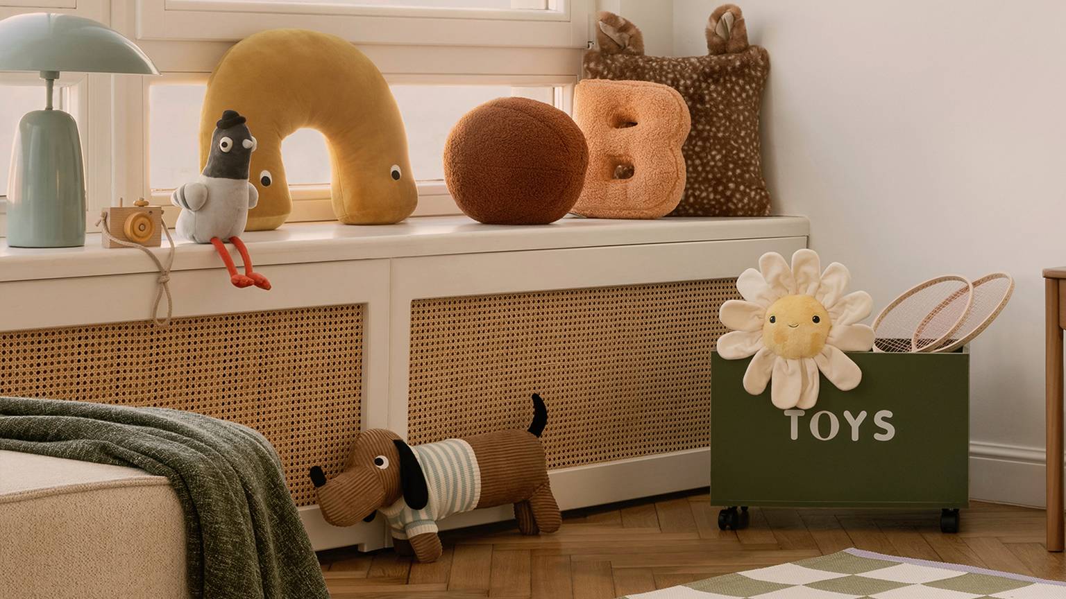 Gemütliches Kinderzimmer mit Plüschtieren, einem Blumenkissen und einer grünen Spielzeugkiste. Sanfte Beleuchtung und ein karierter Teppich sorgen für eine warme Atmosphäre.
