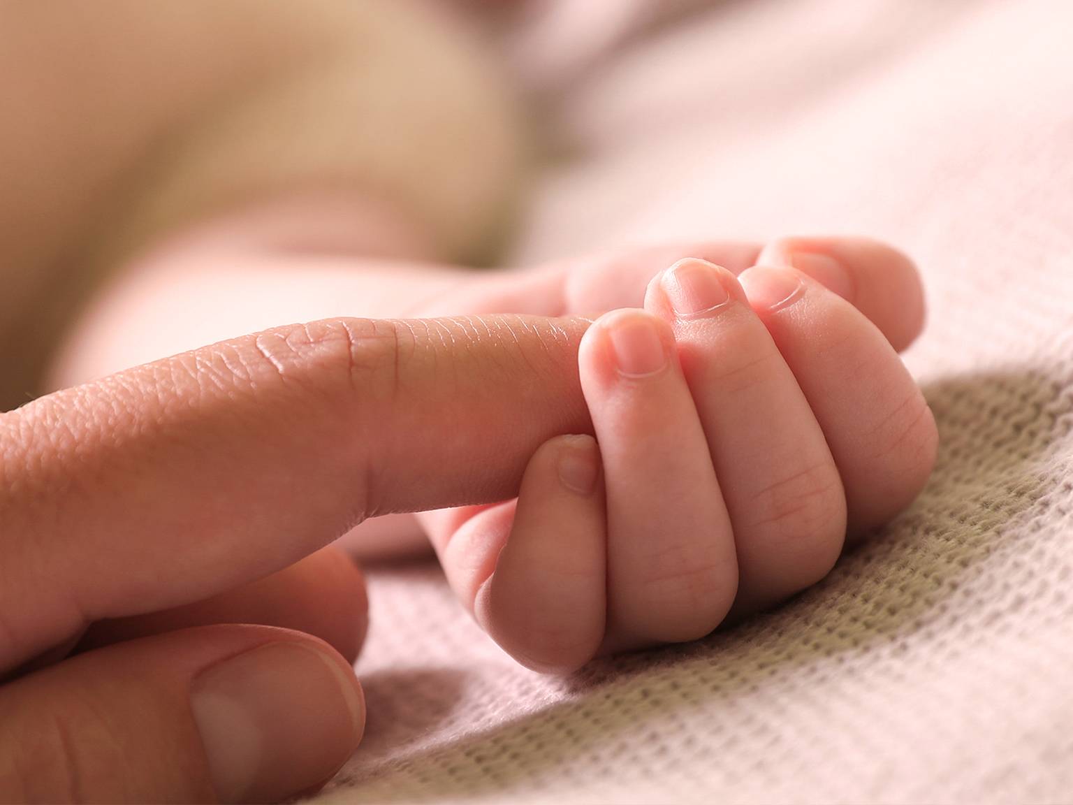 Nahaufnahme einer Babyhand, die sanft den Finger eines Erwachsenen hält und auf einer weichen, beigen Stoffoberfläche ruht.