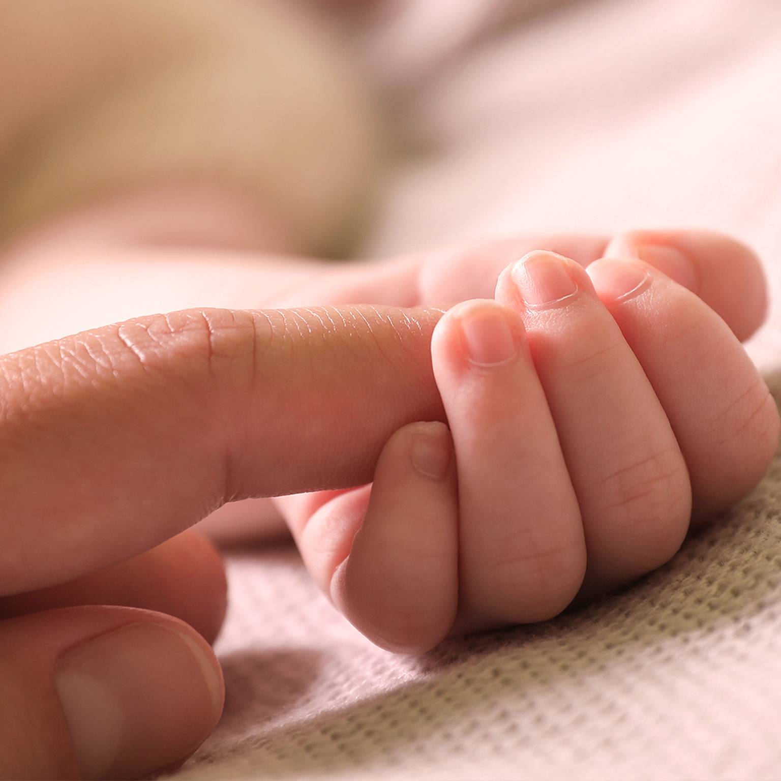 Nahaufnahme einer Babyhand, die sanft den Finger eines Erwachsenen hält und auf einer weichen, beigen Stoffoberfläche ruht.