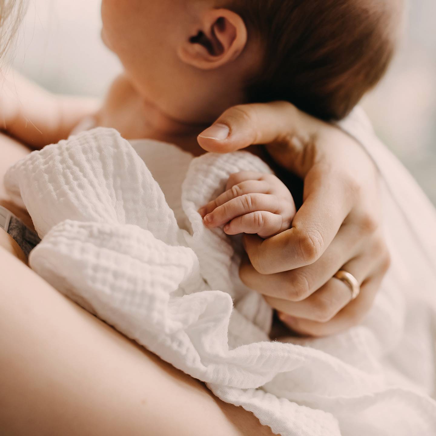 Ein Baby ruht auf der Schulter einer Person, ist in eine weiße Decke gehüllt und wird von der Person sanft an der Hand gehalten.