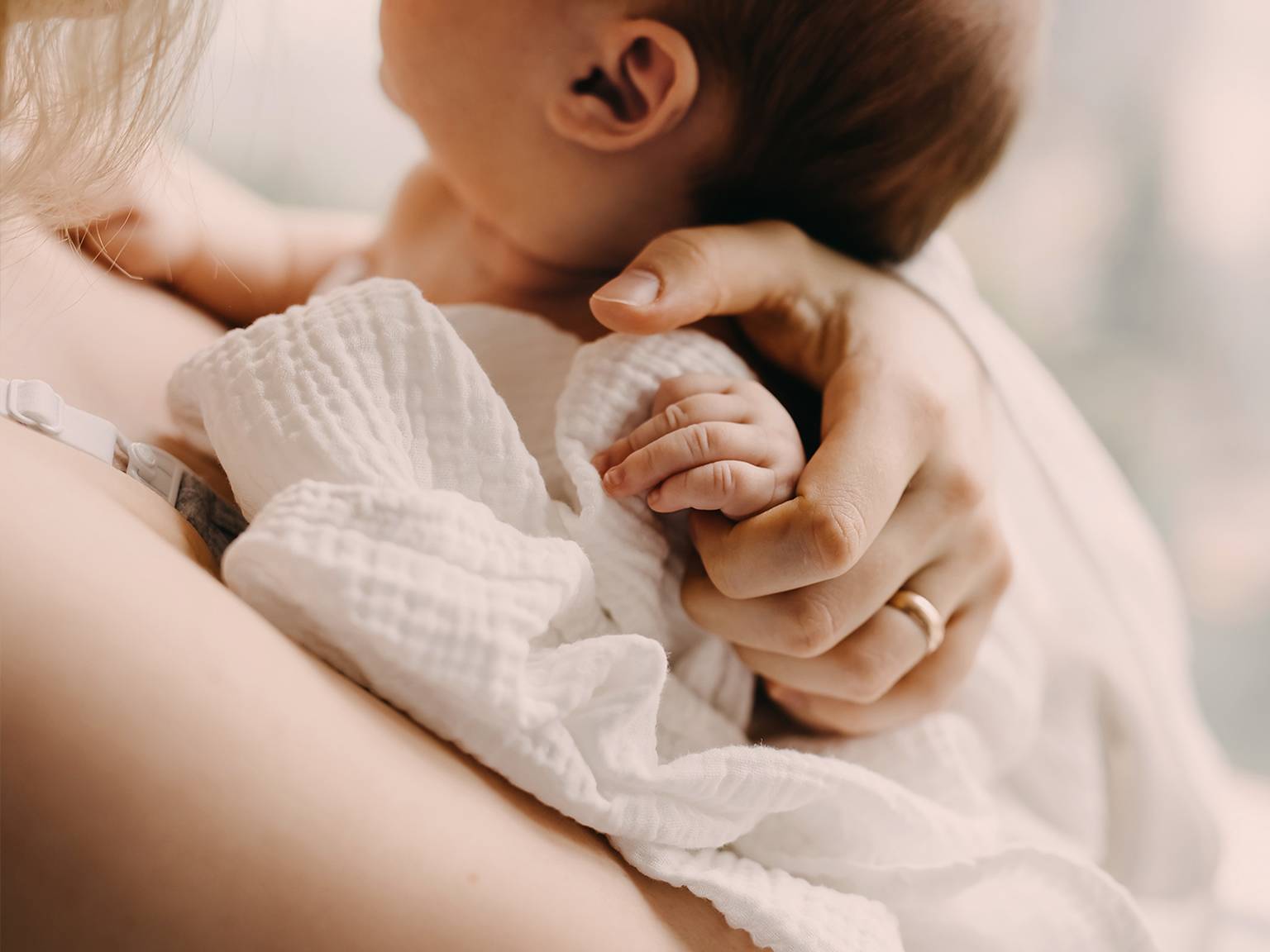 Ein Baby ruht auf der Schulter einer Person, ist in eine weiße Decke gehüllt und wird von der Person sanft an der Hand gehalten.