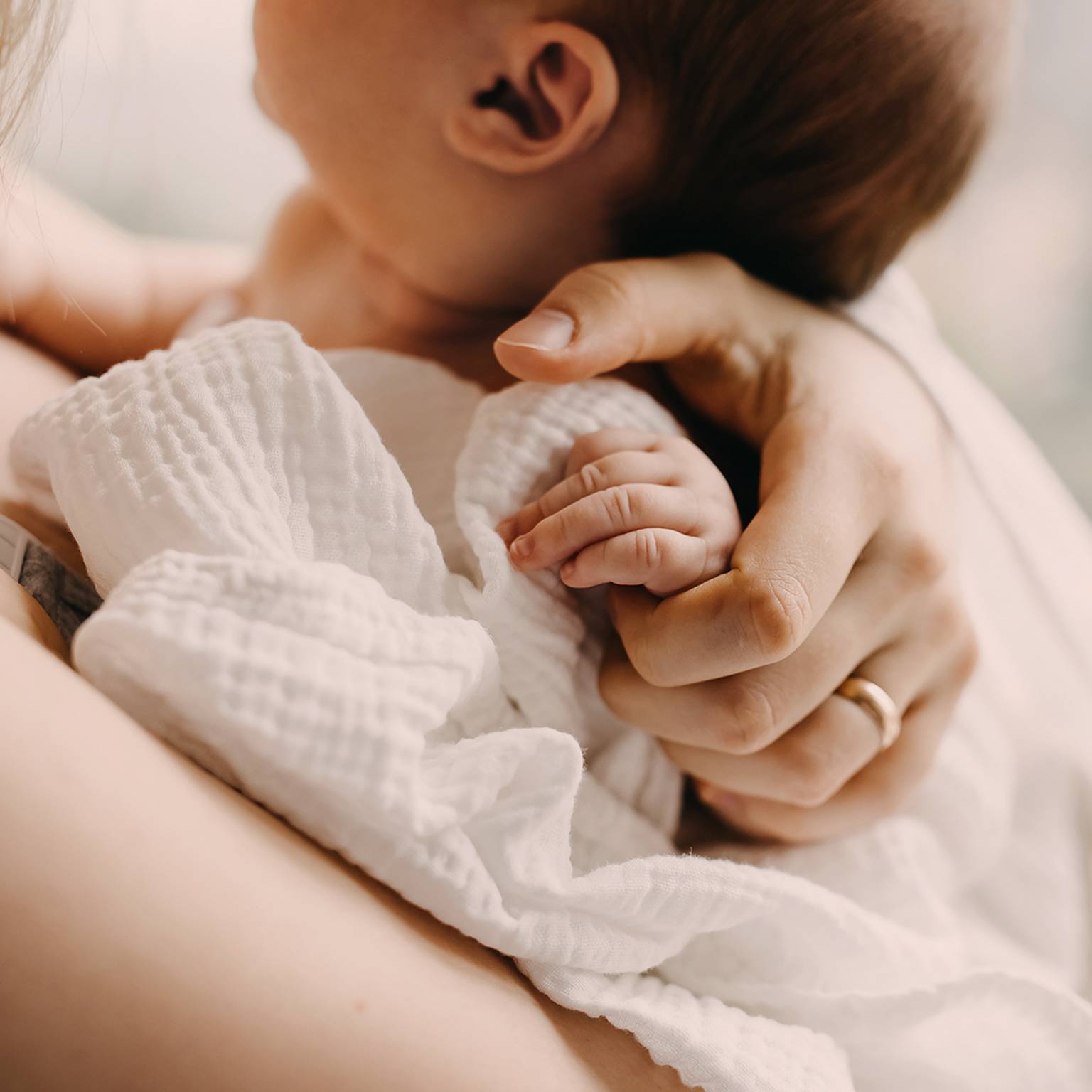 Ein Baby ruht auf der Schulter einer Person, ist in eine weiße Decke gehüllt und wird von der Person sanft an der Hand gehalten.