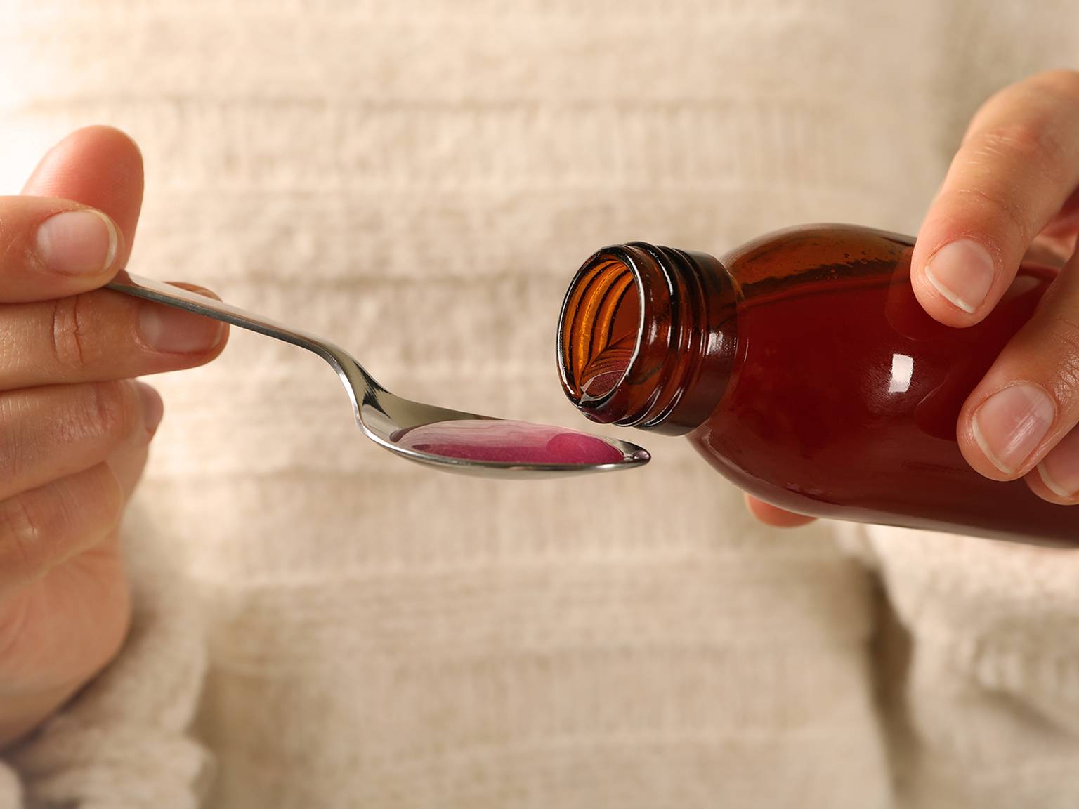 Vor einem weichen, strukturierten Hintergrund gießt eine Person rosafarbene Medikamentenflüssigkeit aus einer braunen Flasche in einen Löffel.