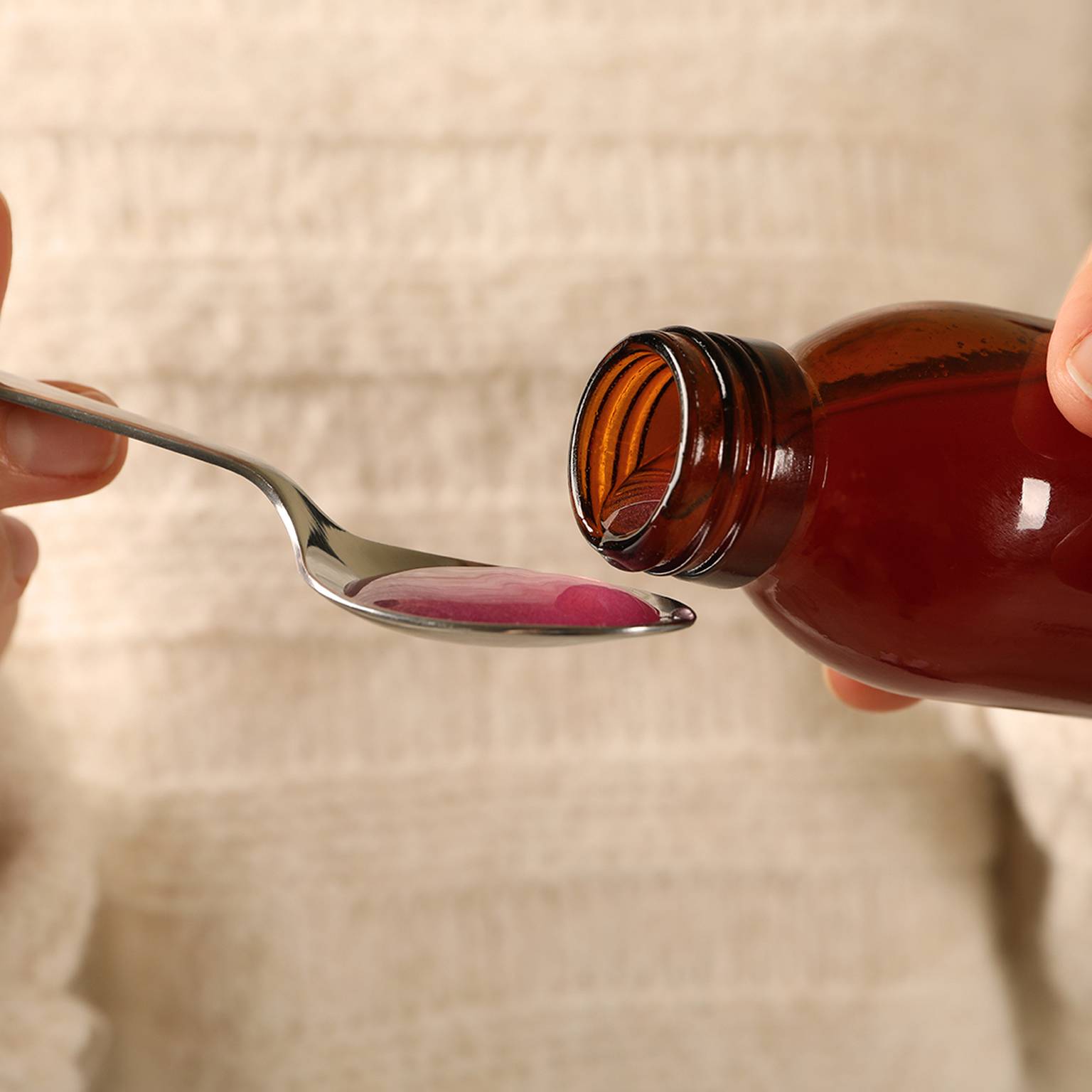 Vor einem weichen, strukturierten Hintergrund gießt eine Person rosafarbene Medikamentenflüssigkeit aus einer braunen Flasche in einen Löffel.