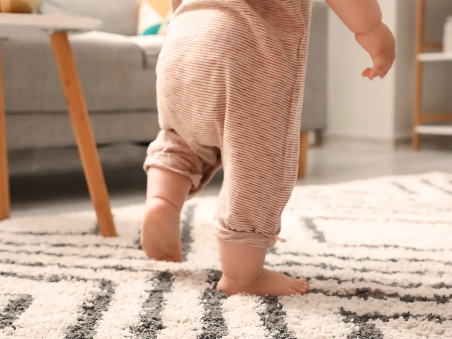 Ein Baby im gestreiften Pyjama macht einen Schritt auf einen gemusterten Teppich in einem gemütlichen Wohnzimmer mit einem grauen Sofa und einem Holztisch.