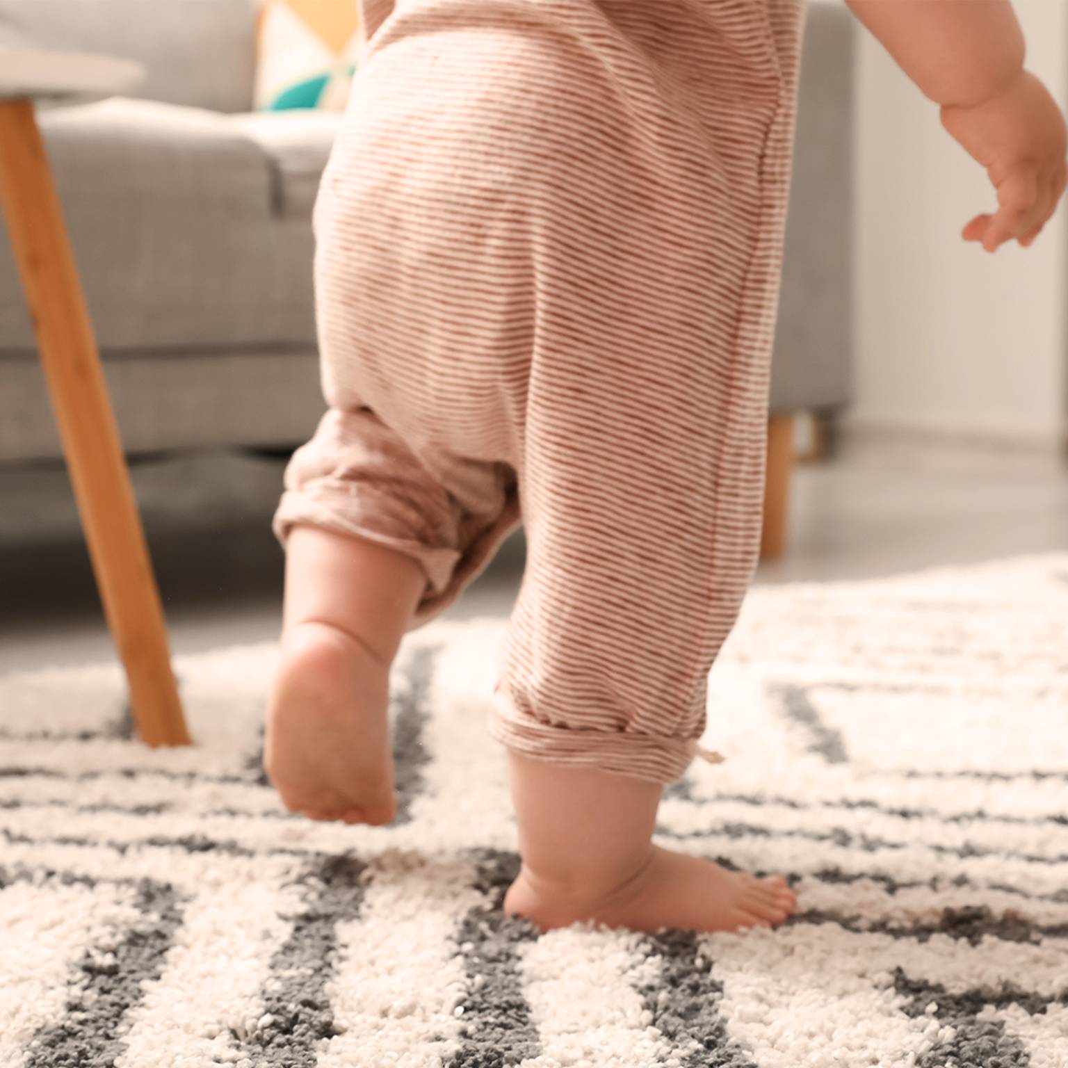Ein Baby im gestreiften Pyjama macht einen Schritt auf einen gemusterten Teppich in einem gemütlichen Wohnzimmer mit einem grauen Sofa und einem Holztisch.