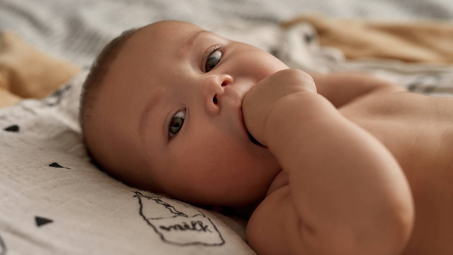 Ein Baby liegt auf einer gemusterten Decke, schaut nach oben und hat eine Hand im Mund.