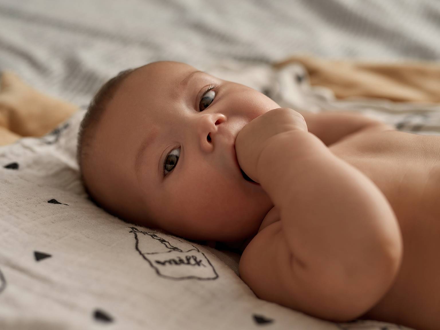 Ein Baby liegt auf einer gemusterten Decke, schaut nach oben und hat eine Hand im Mund.