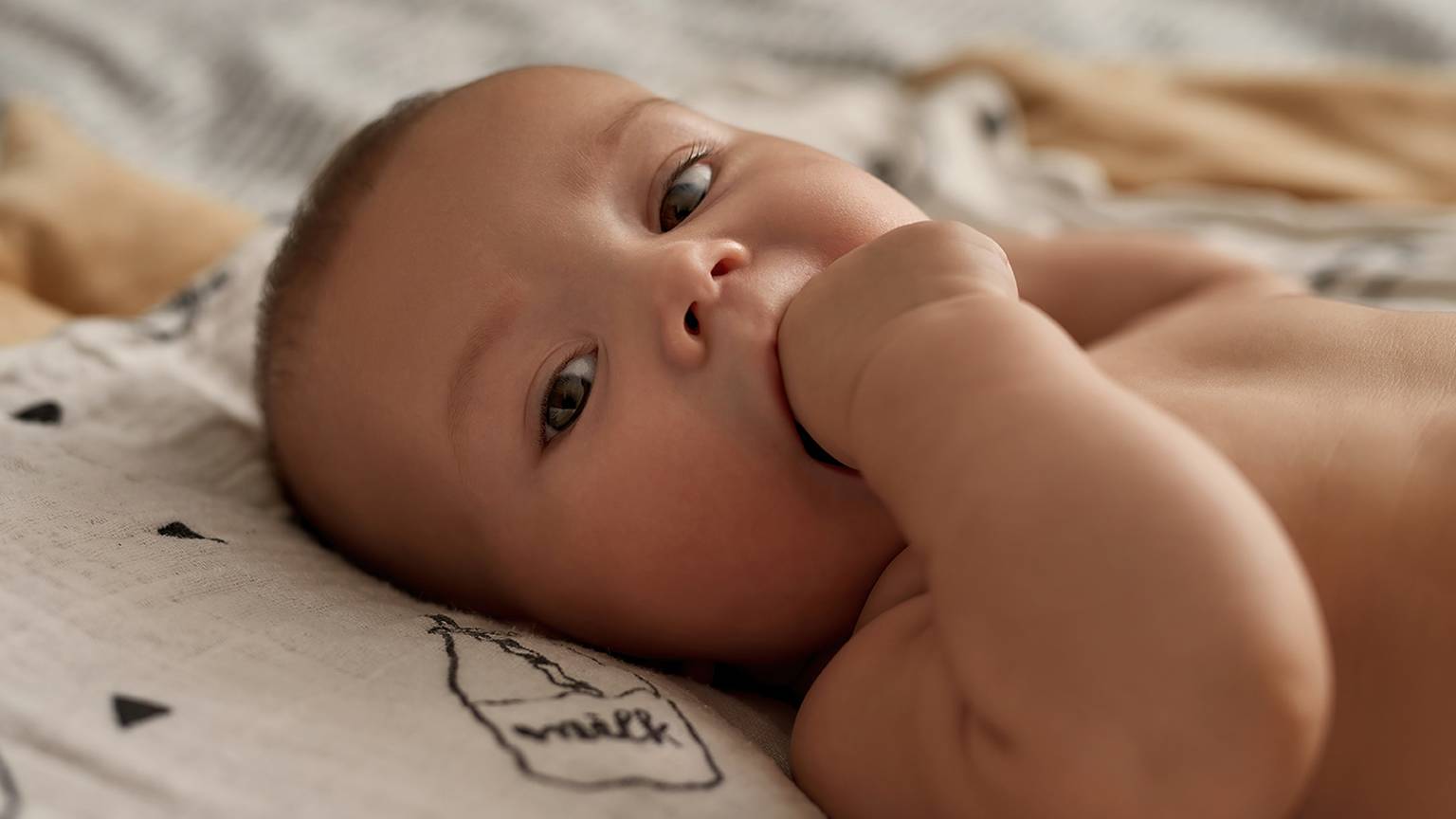 Ein Baby liegt auf einer gemusterten Decke, schaut nach oben und hat eine Hand im Mund.