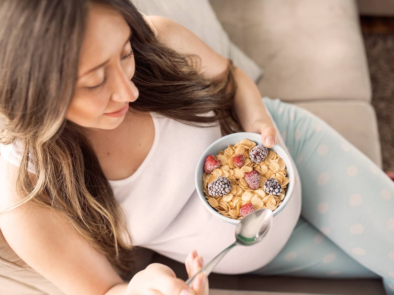 Eine Frau in einem weißen Tanktop und gepunkteter Hose sitzt auf einem Sofa und hält eine Schüssel Müsli mit Brombeeren und Himbeeren in der Hand.
