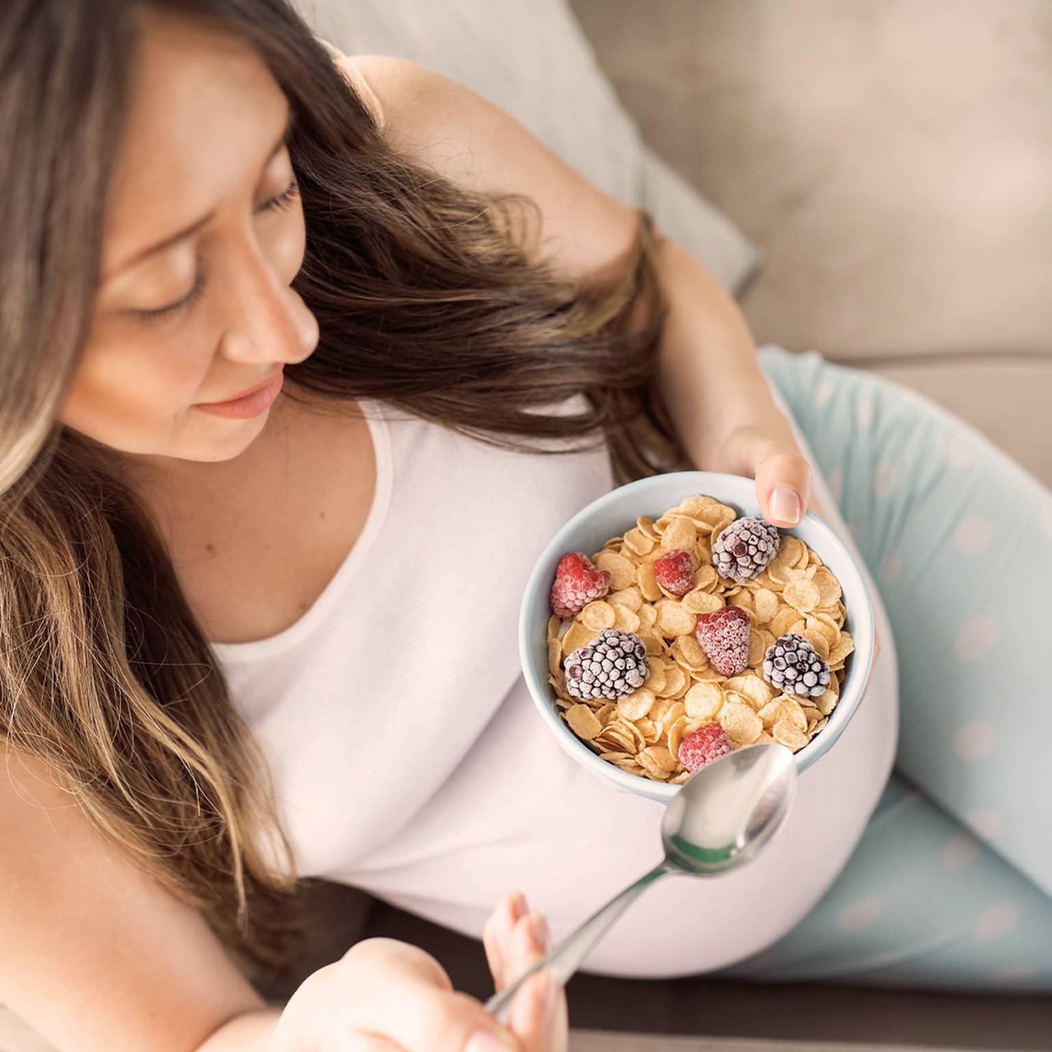 Eine Frau in einem weißen Tanktop und gepunkteter Hose sitzt auf einem Sofa und hält eine Schüssel Müsli mit Brombeeren und Himbeeren in der Hand.