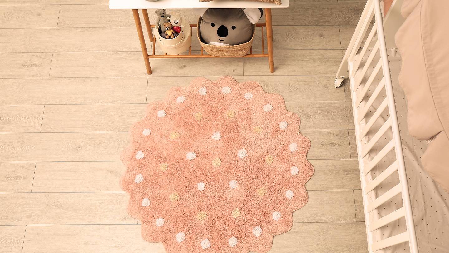 Ein gemütliches Kinderzimmer mit einem rosafarbenen Teppich mit weißen und gelben Punkten, einem weißen Kinderbett, einem Tisch mit einer Lampe und Stofftieren.