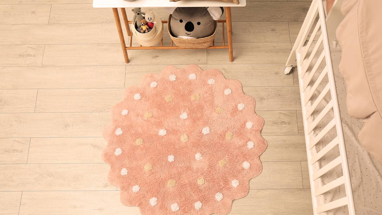 Ein gemütliches Kinderzimmer mit einem rosafarbenen Teppich mit weißen und gelben Punkten, einem weißen Kinderbett, einem Tisch mit einer Lampe und Stofftieren.