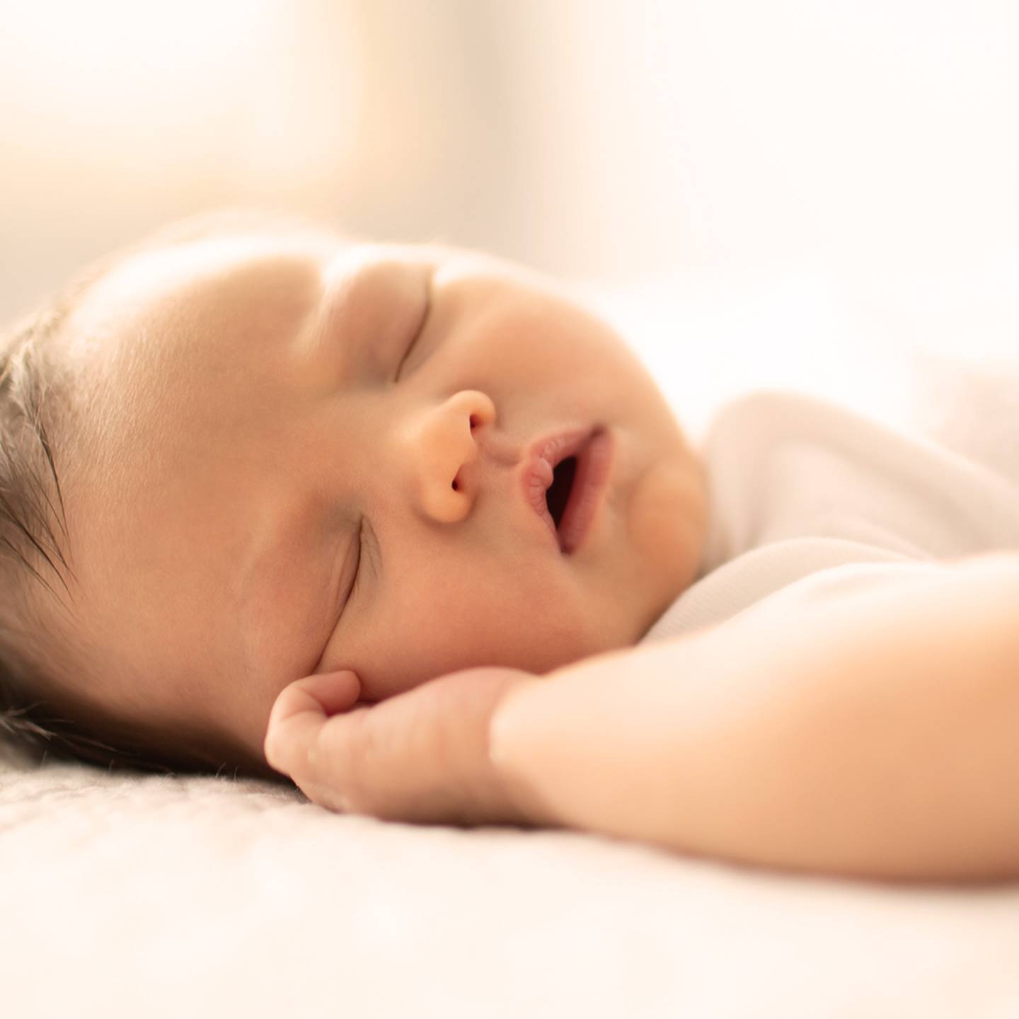 Ein schlafendes Baby liegt friedlich auf einer weichen Oberfläche und trägt ein weißes Outfit. Die warme und sanfte Beleuchtung schafft eine ruhige Atmosphäre.