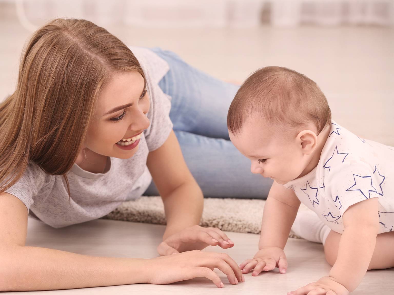 Eine lächelnde Frau liegt auf dem Boden und beschäftigt sich mit einem krabbelnden Baby in einem sterngemusterten Strampler auf einer hellen Fläche.