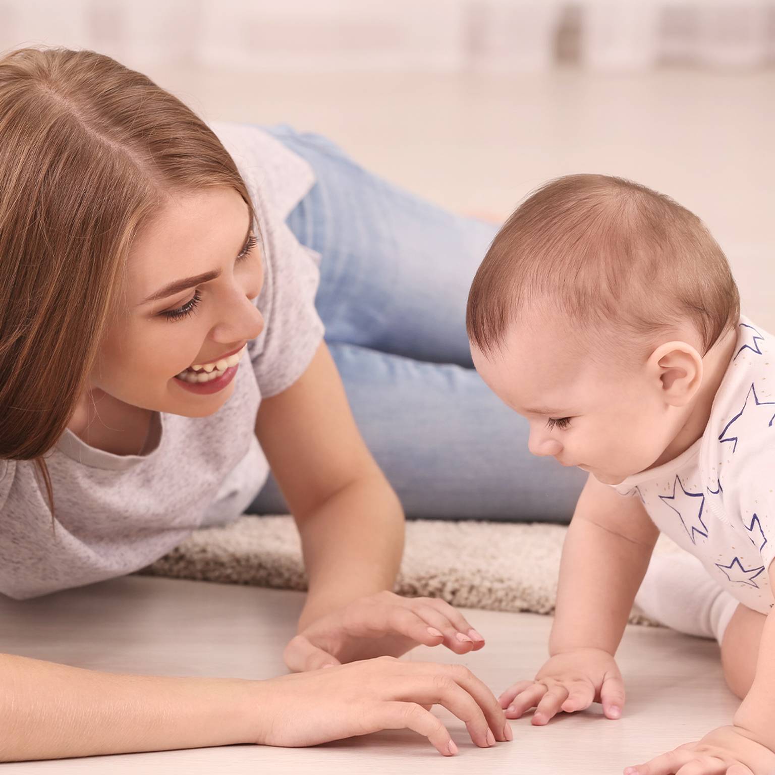 Eine lächelnde Frau liegt auf dem Boden und beschäftigt sich mit einem krabbelnden Baby in einem sterngemusterten Strampler auf einer hellen Fläche.