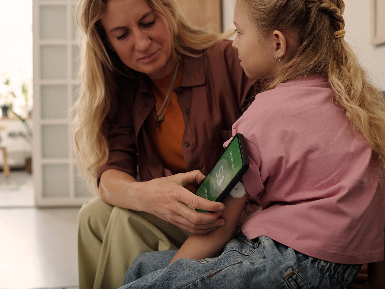 Eine Frau misst den Blutzucker eines jungen Mädchens mithilfe einer Smartphone-App. Am Arm des Mädchens befindet sich ein Sensor. Die beiden sitzen im Haus.