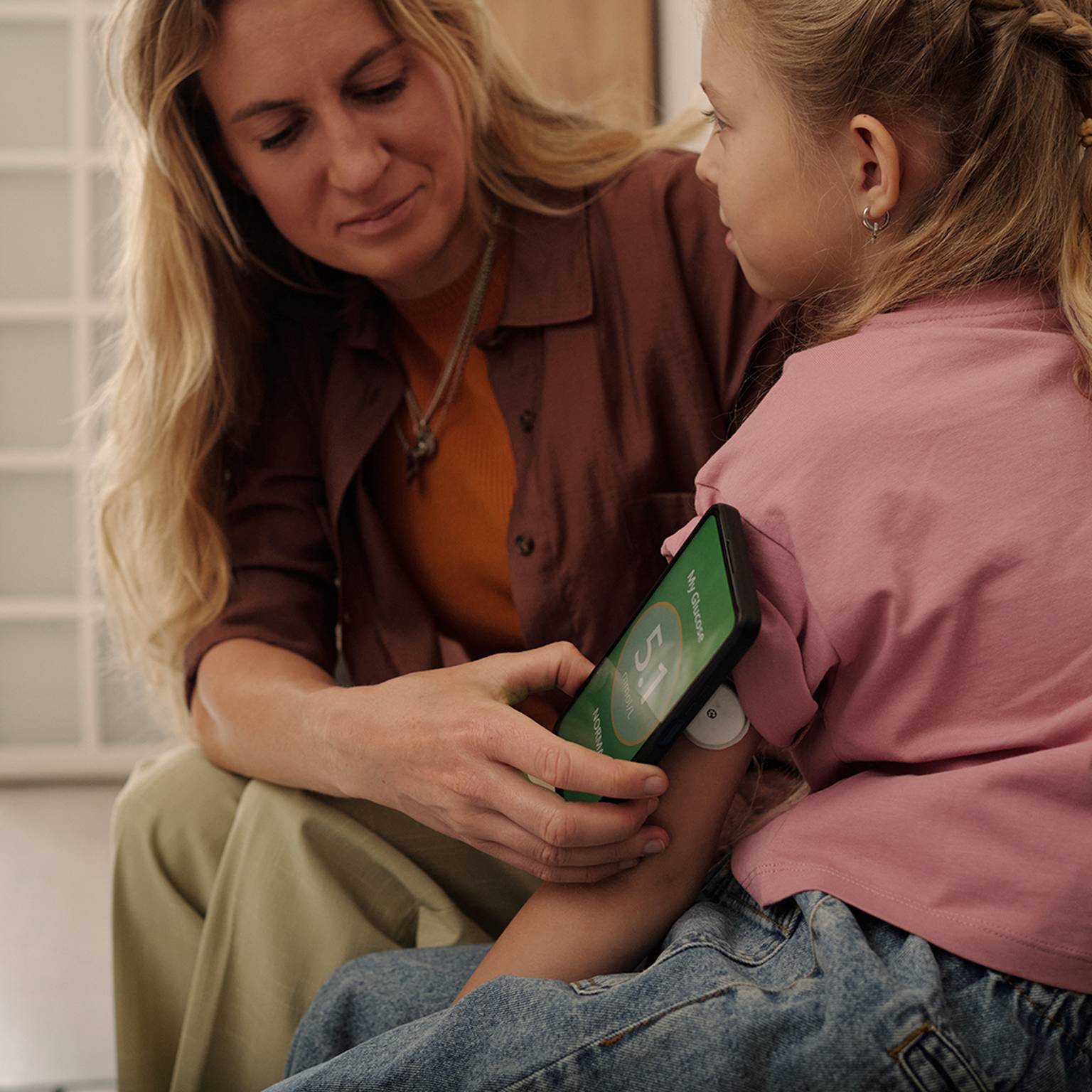 Eine Frau misst den Blutzucker eines jungen Mädchens mithilfe einer Smartphone-App. Am Arm des Mädchens befindet sich ein Sensor. Die beiden sitzen im Haus.