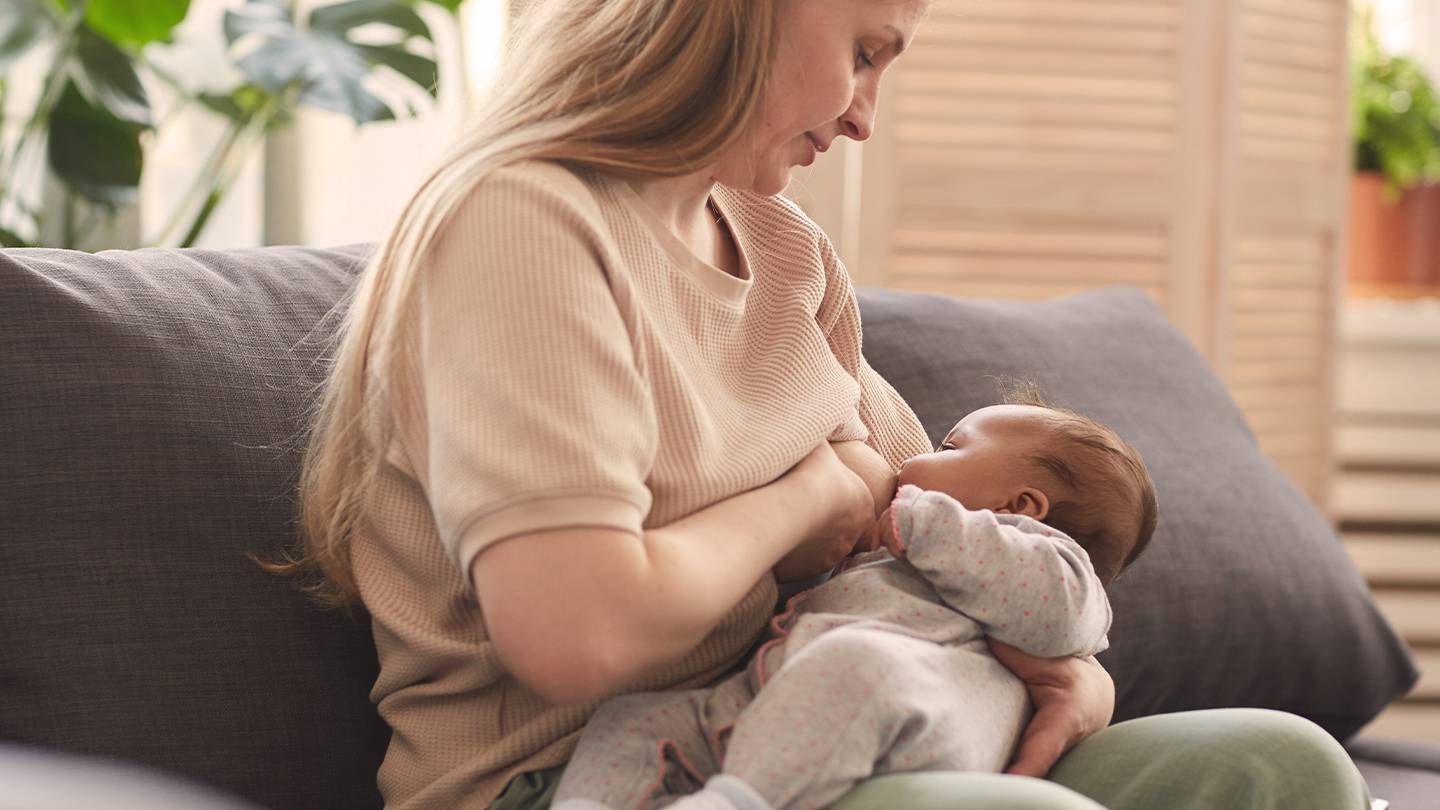 Eine Frau mit langen Haaren stillt ein Baby auf einem Sofa, umgeben von Pflanzen und einem sanft beleuchteten Raum.