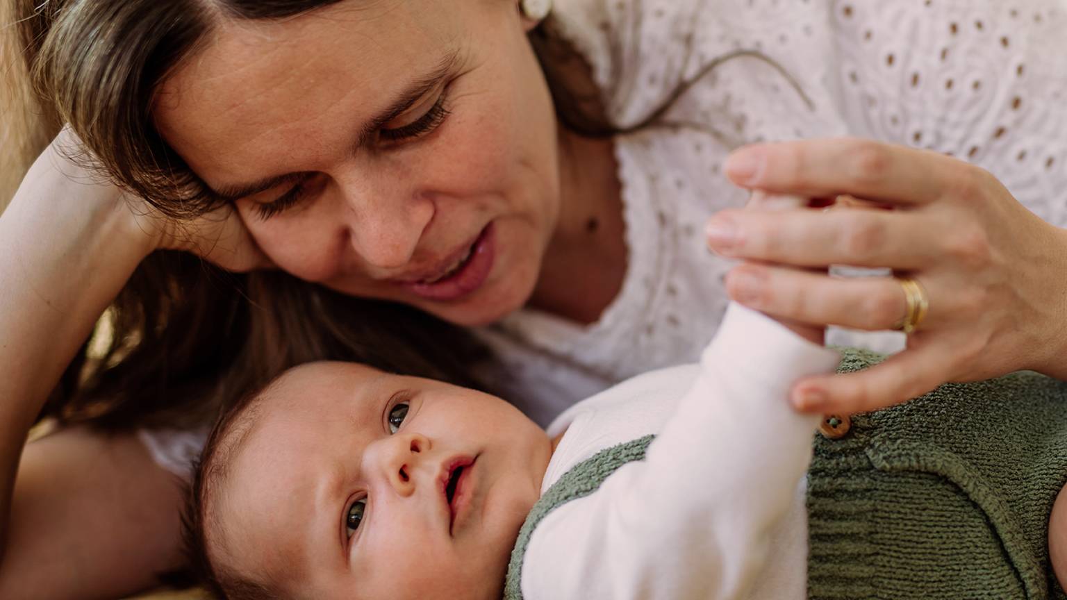 Eine Frau und ein Baby liegen auf einer weichen Unterlage. Die Frau hält sanft die Hand des Babys. Beide sehen zufrieden und entspannt aus.