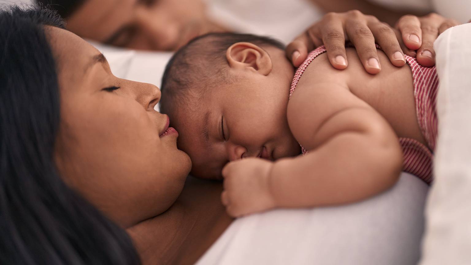 Eine Frau schläft friedlich mit einem Baby auf ihrer Brust, während im Hintergrund eine andere Person schläft.