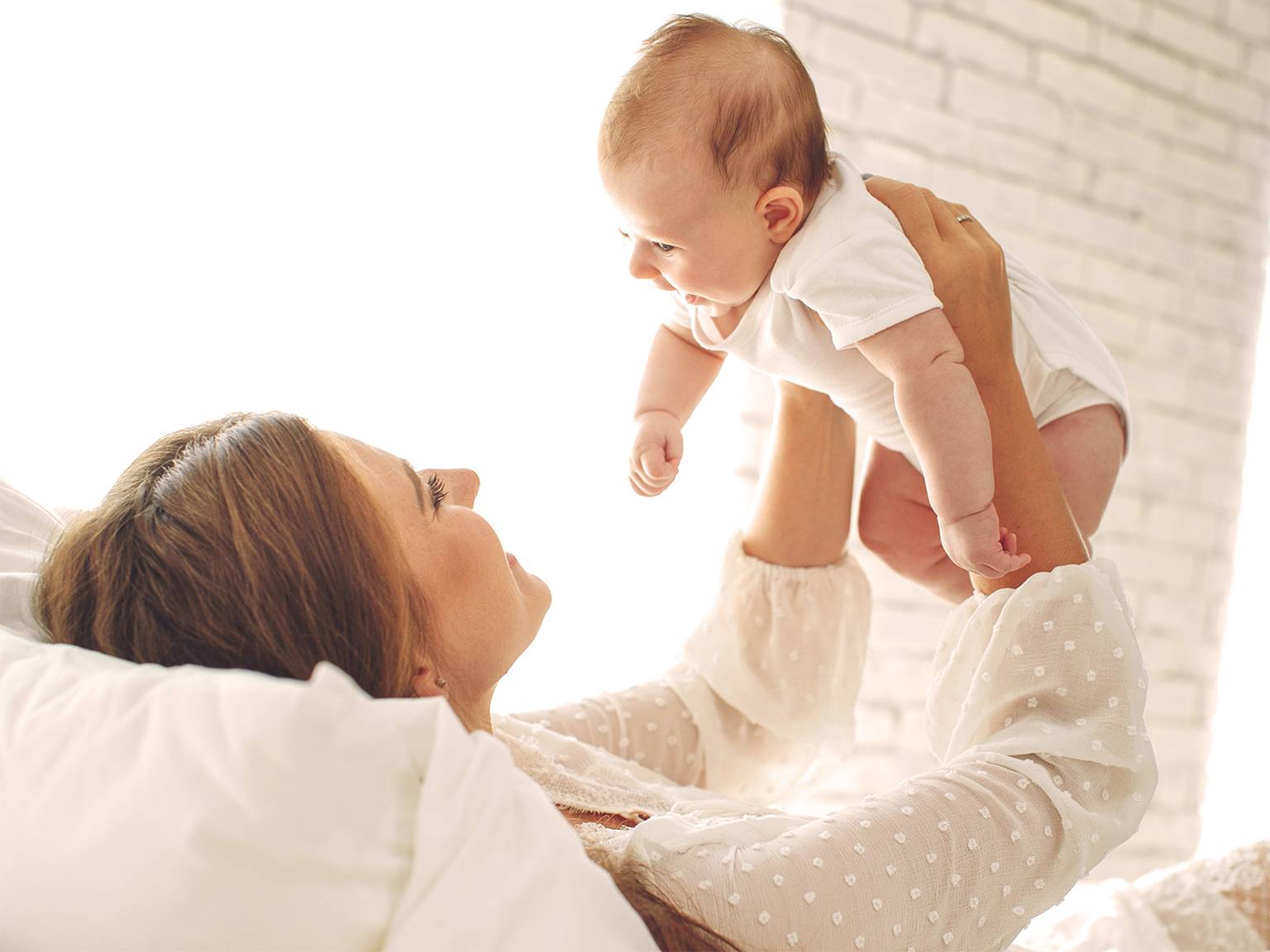 Eine Frau liegt auf einem Bett und hebt freudig ein Baby in einem weißen Strampler hoch. Sie befinden sich in einem hellen Raum mit einer weißen Ziegelwand im Hintergrund.