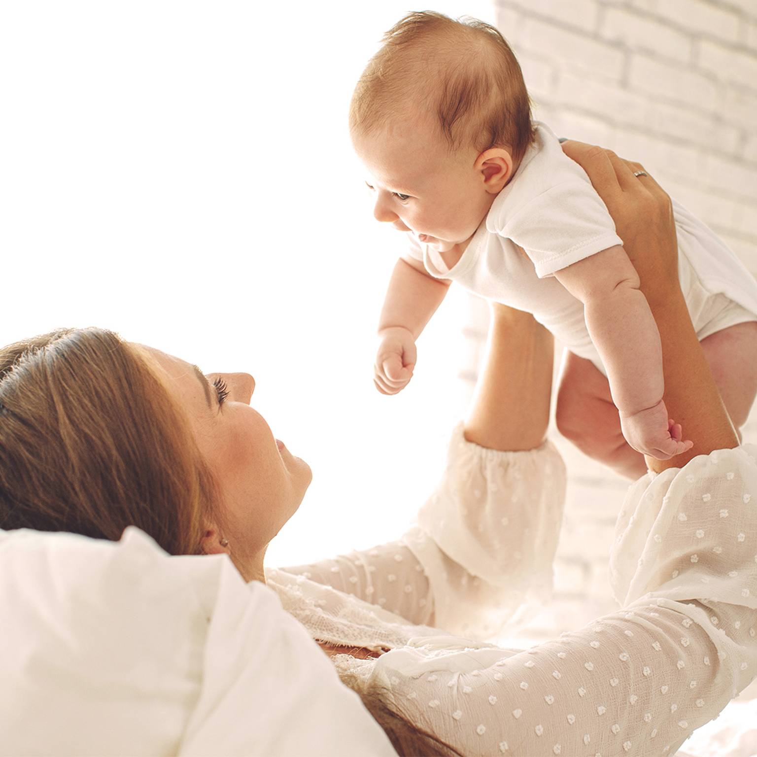 Eine Frau liegt auf einem Bett und hebt freudig ein Baby in einem weißen Strampler hoch. Sie befinden sich in einem hellen Raum mit einer weißen Ziegelwand im Hintergrund.