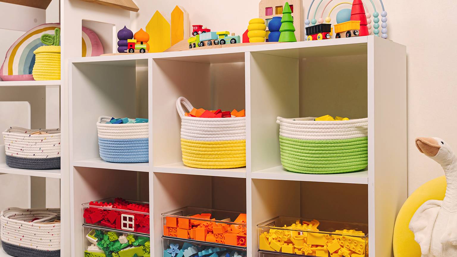 Ein farbenfrohes Kinderzimmerregal mit Körben voller Spielzeug, Blöcken und Spielzeugeisenbahnen, mit Gänseplüsch und Regenbogendekor.