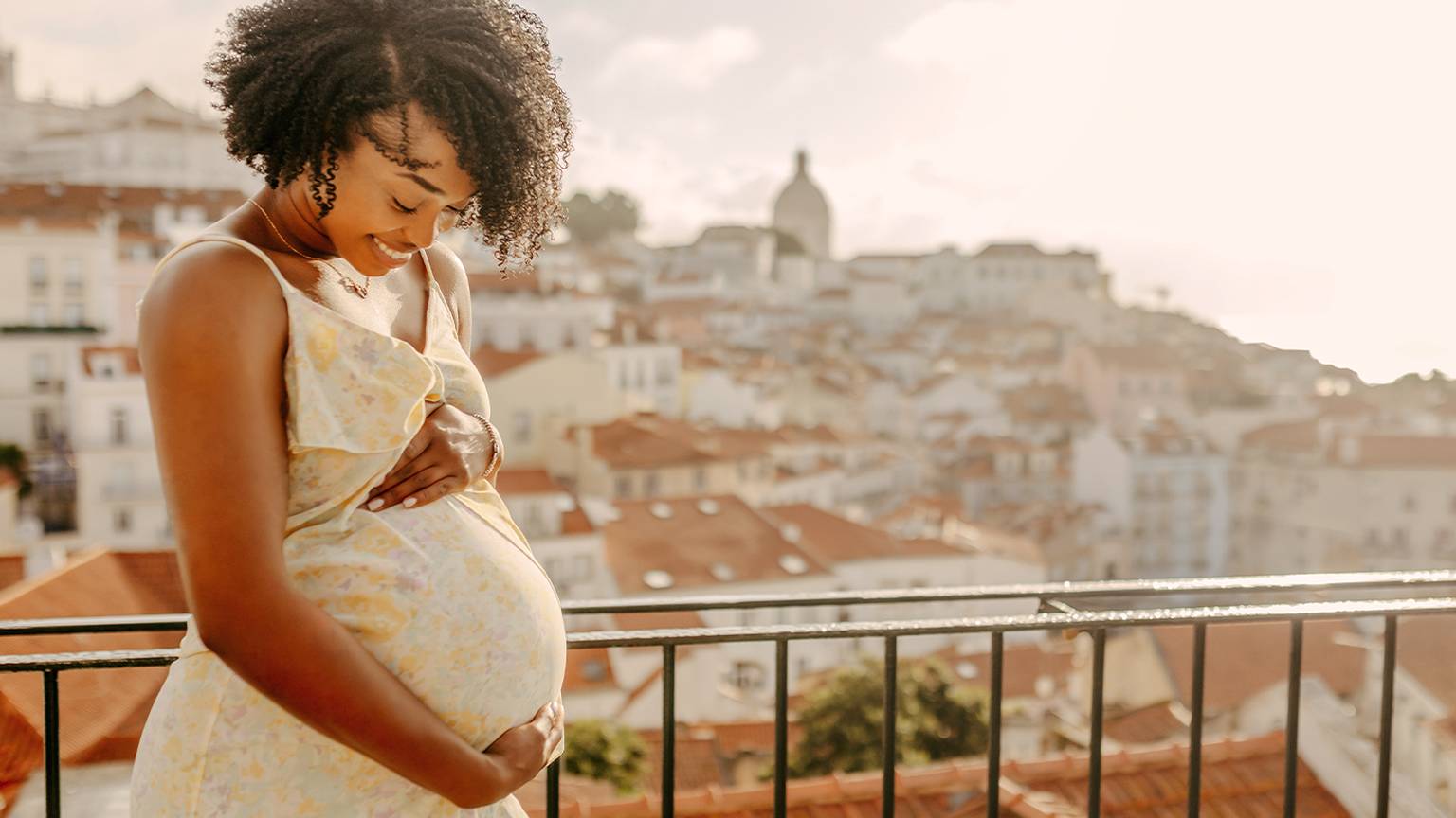 Eine schwangere Frau in einem geblümten Kleid lächelt und hält ihren Bauch auf einem Balkon mit Blick auf eine Stadtlandschaft mit Terrakotta-Dächern.
