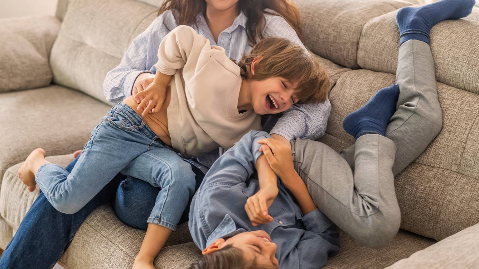 Eine Frau kitzelt spielerisch zwei lachende Kinder auf einer Couch und schafft so eine fröhliche und lebhafte Atmosphäre.