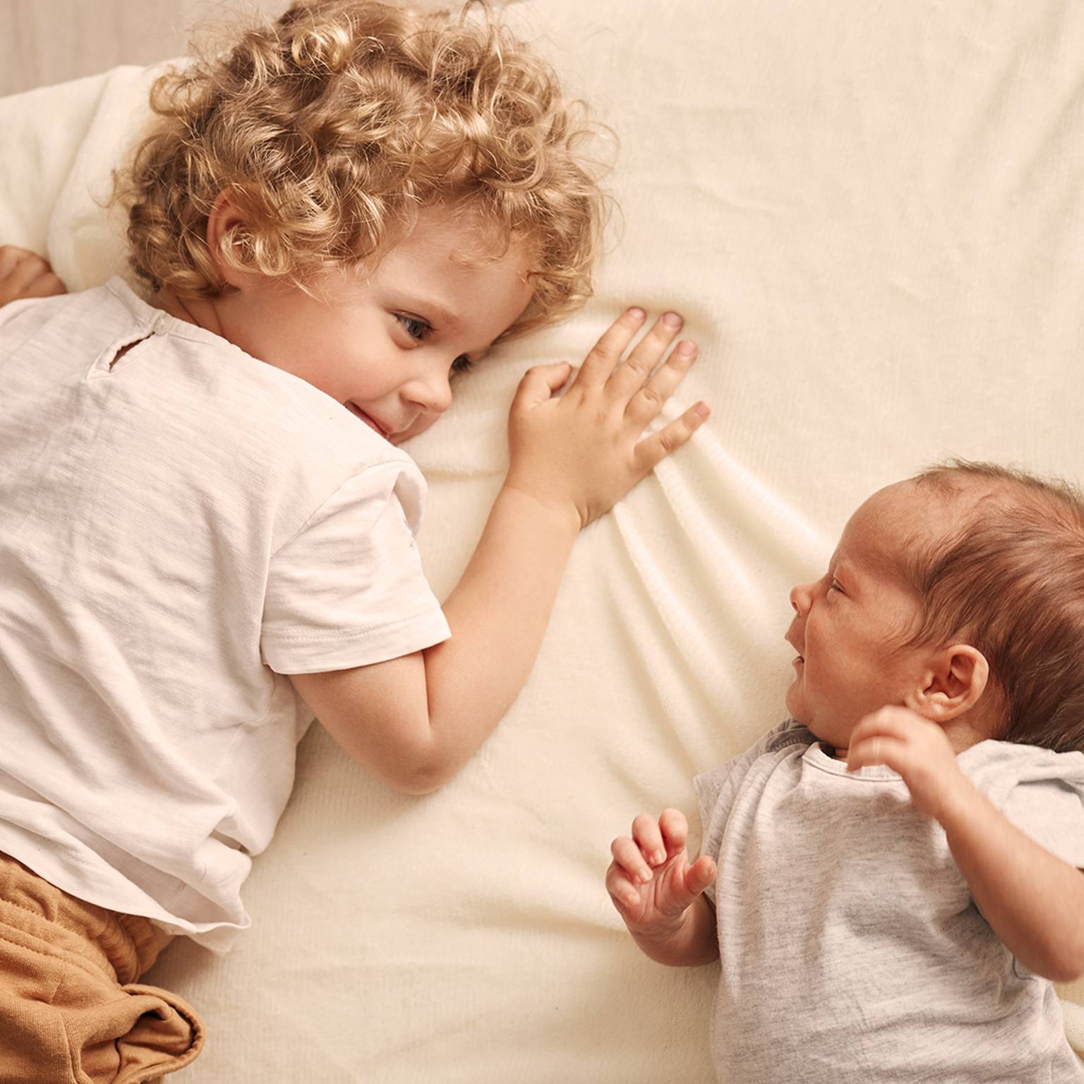 Ein kleines Kind mit lockigem Haar liegt auf einem Bett und lächelt ein neugeborenes Baby an, das neben ihm liegt. Beide tragen helle Kleidung.