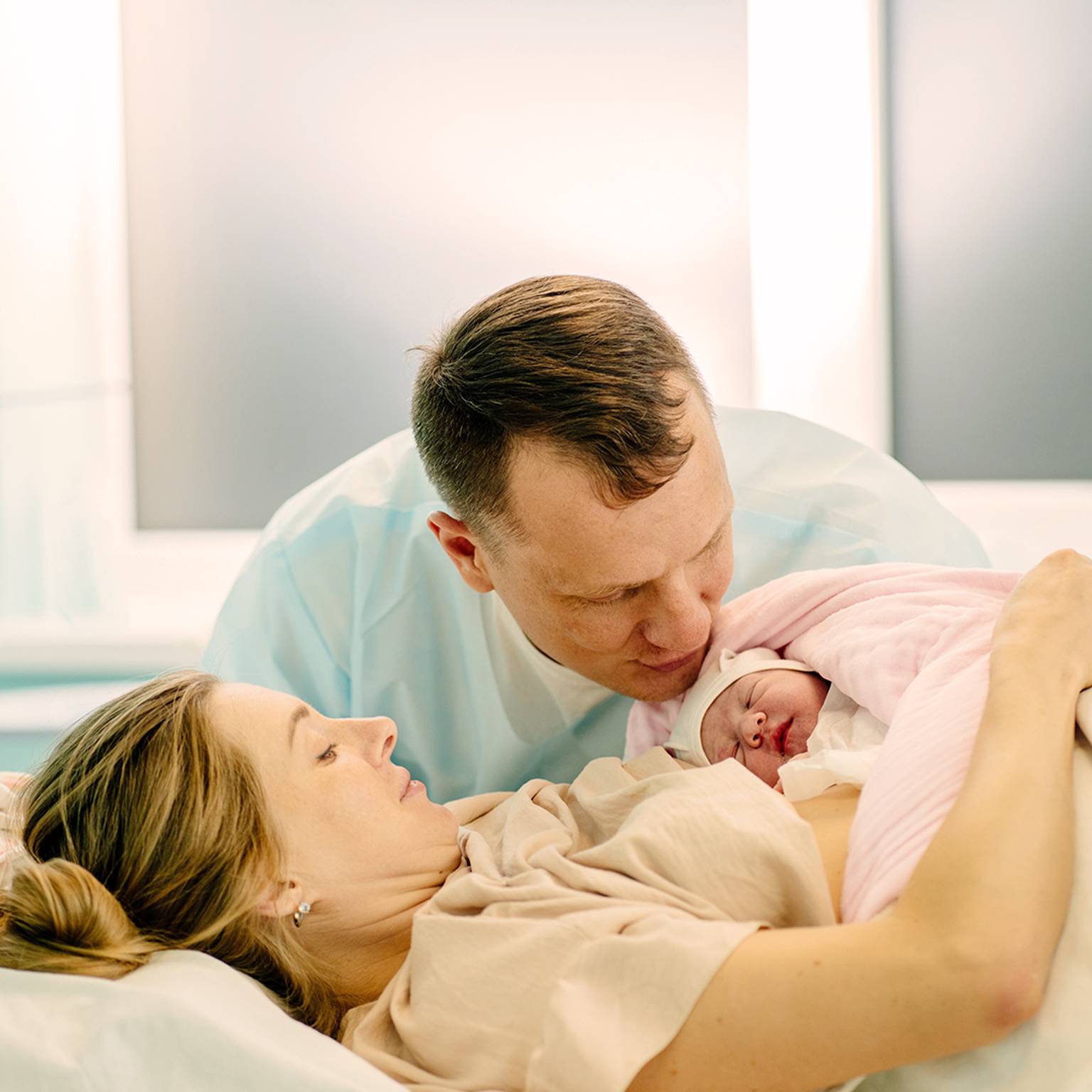 Eine Mutter liegt in einem Krankenhausbett und hält ein in eine rosa Decke gewickeltes Neugeborenes, während der Vater sanft den Kopf des Babys küsst.