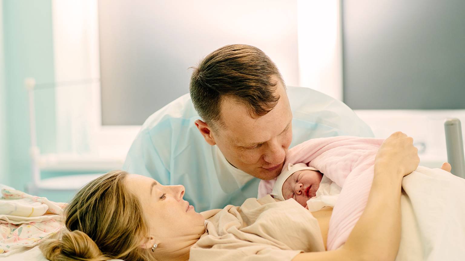 Eine Mutter liegt in einem Krankenhausbett und hält ein in eine rosa Decke gewickeltes Neugeborenes, während der Vater sanft den Kopf des Babys küsst.