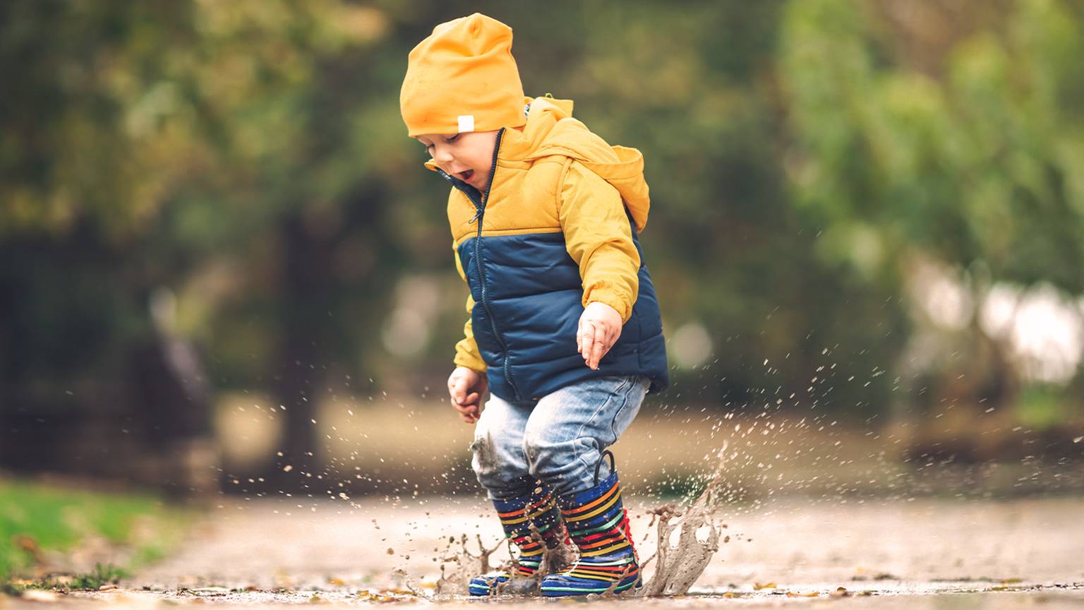 Ein Kind mit gelber Mütze und Jacke planscht fröhlich in einer Pfütze, trägt bunte Gummistiefel, im Hintergrund sind verschwommene grüne Bäume zu sehen.