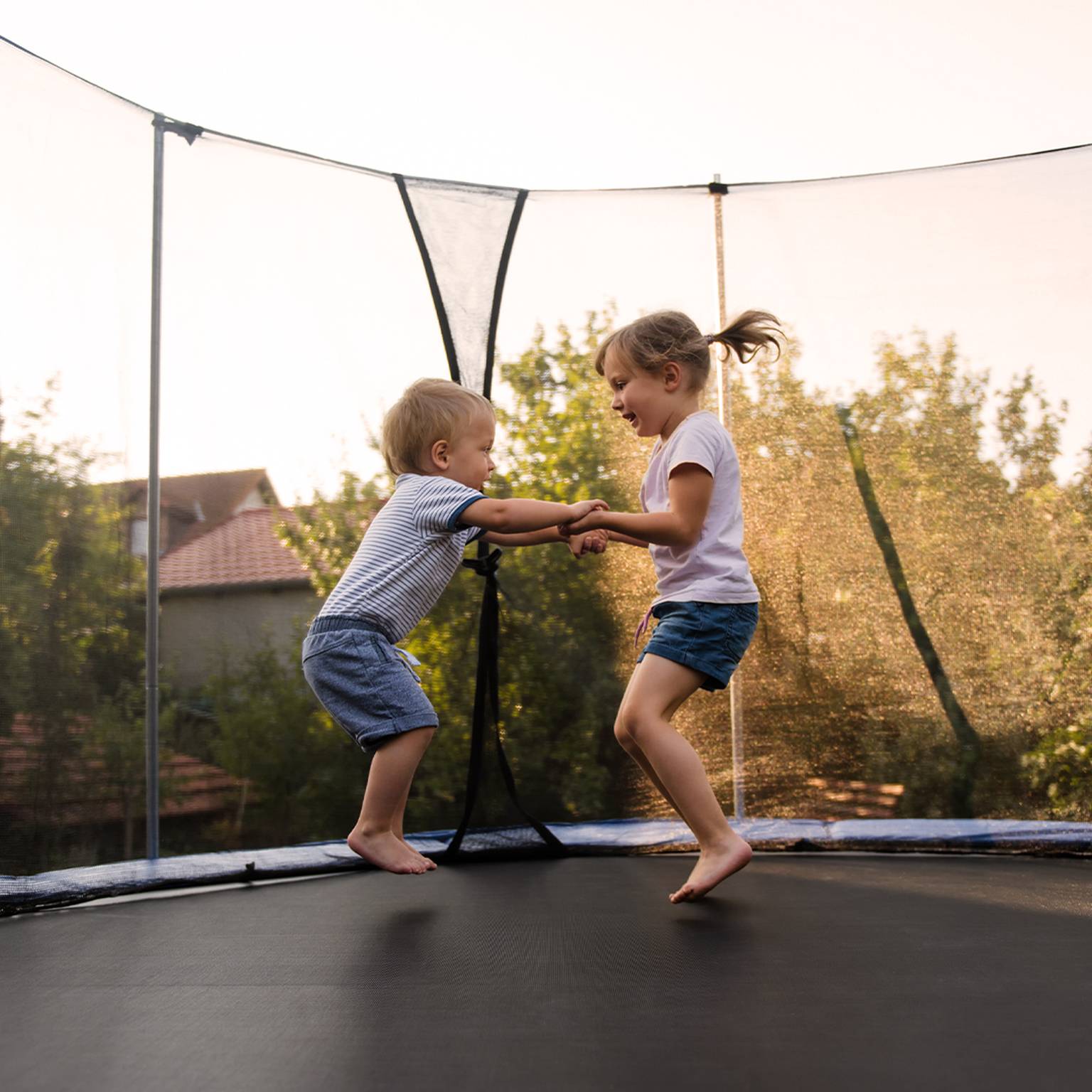 Zwei kleine Kinder spielen fröhlich Händchen haltend auf einem Trampolin, im Hintergrund sind Bäume und Häuser unter einem klaren Himmel zu sehen.