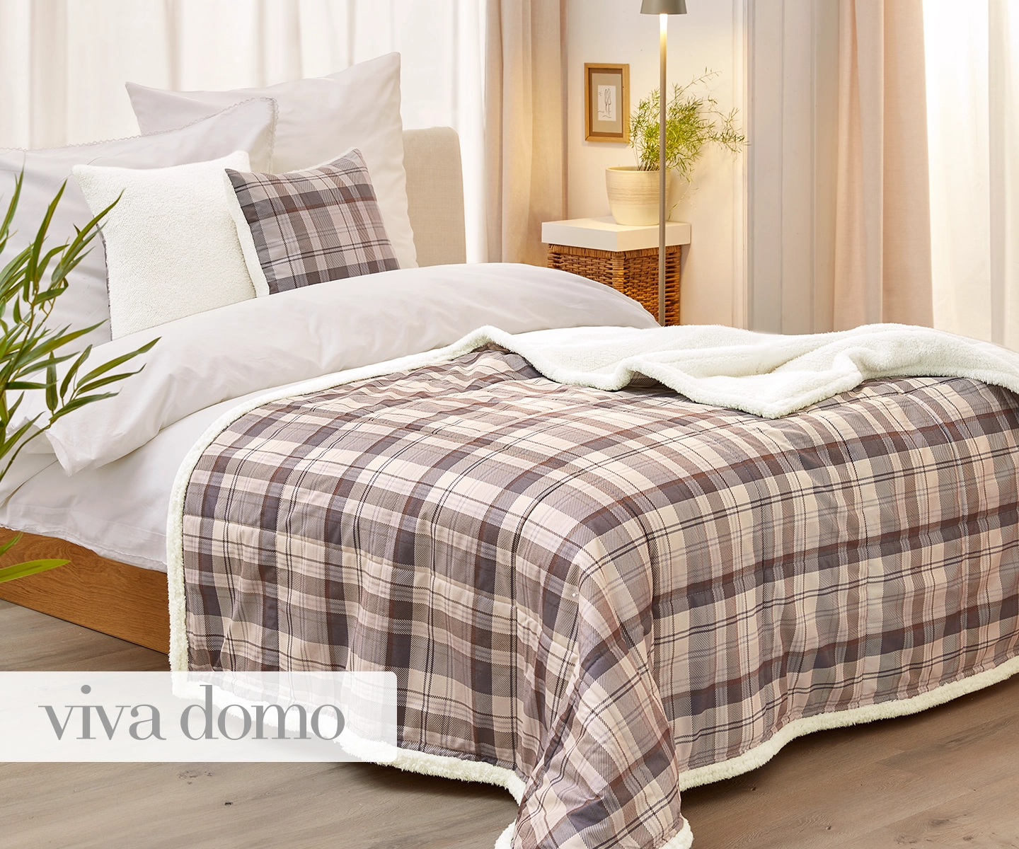 Gemütliches Schlafzimmer mit einer karierten Tagesdecke, weißen Kissen, einem Nachttisch mit Lampe und einer Pflanze. Sanftes Tageslicht erfüllt den Raum.