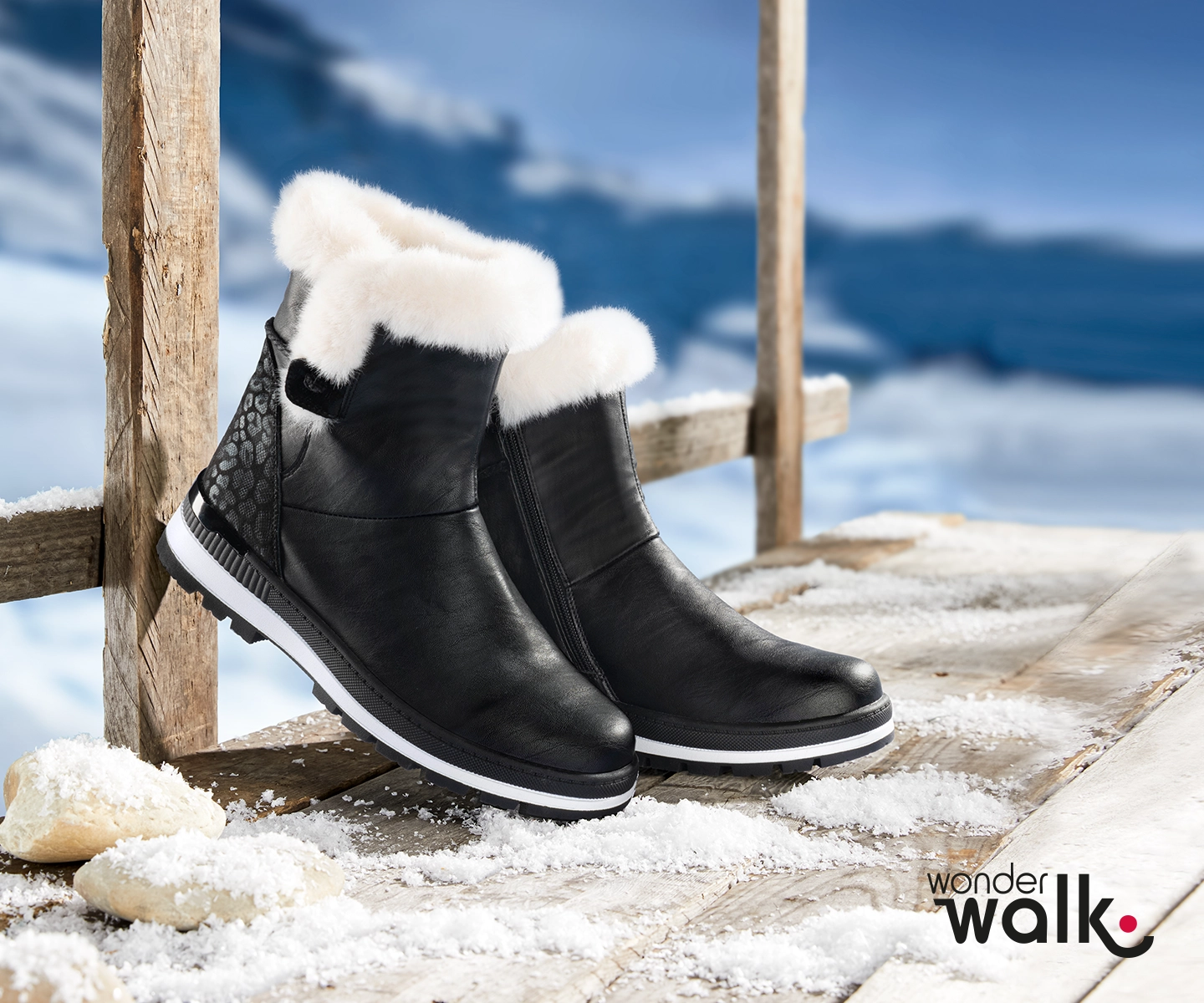 Schwarze Winterstiefel mit weißem Fellfutter stehen auf einem schneebedeckten Holzsteg, im Hintergrund eine verschneite Berglandschaft.