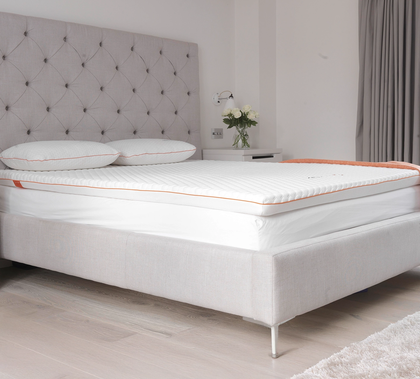 Ein helles Schlafzimmer mit einem ordentlich gemachten Bett, darauf ein weißer Matratzentopper, weiße Bettwäsche, eine orangefarbenen Tagesdecke, zwei Kissen. Daneben ein Beistelltisch mit Blumen.