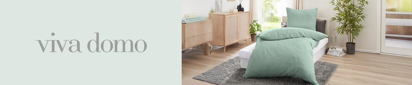 Schlafzimmer mit hellgrüner Bettwäsche, einer Kommode aus Holz und einer Topfpflanze. Helle und luftige Atmosphäre mit sanftem Tageslicht. Links der Schriftzug „Viva Domo”.