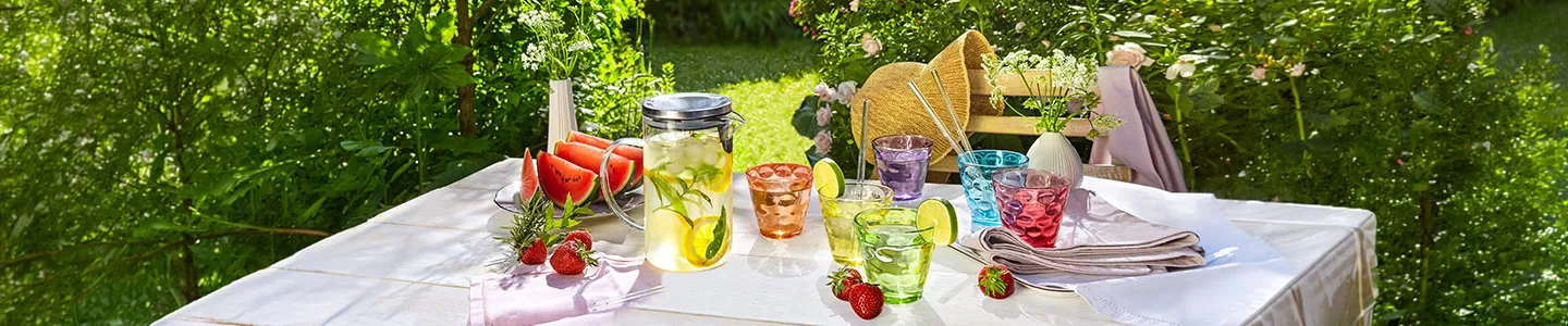 Ein sonniger Gartentisch, gedeckt mit bunten Gläsern, einem Krug Zitronenwasser, frischen Wassermelonenscheiben, Erdbeeren und Sonnenhüten auf Stühlen, umgeben von Grün.