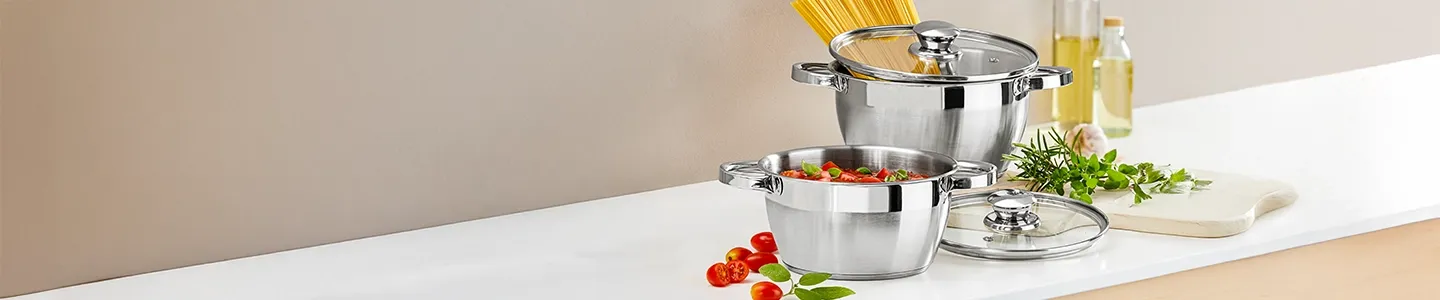 Zwei Edelstahltöpfe auf einer Küchenarbeitsplatte, in dem einen Spaghetti, in dem anderen Tomaten. Daneben liegen Kräuter, Öl und Essig.