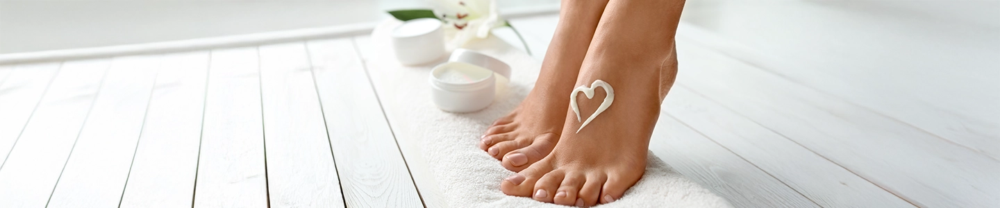 Nahaufnahme von Füßen auf einem weißen Handtuch mit einem herzförmigen Crememuster auf der Haut. Im Hintergrund stehen auf einem Holzboden geöffnete Cremetiegel.