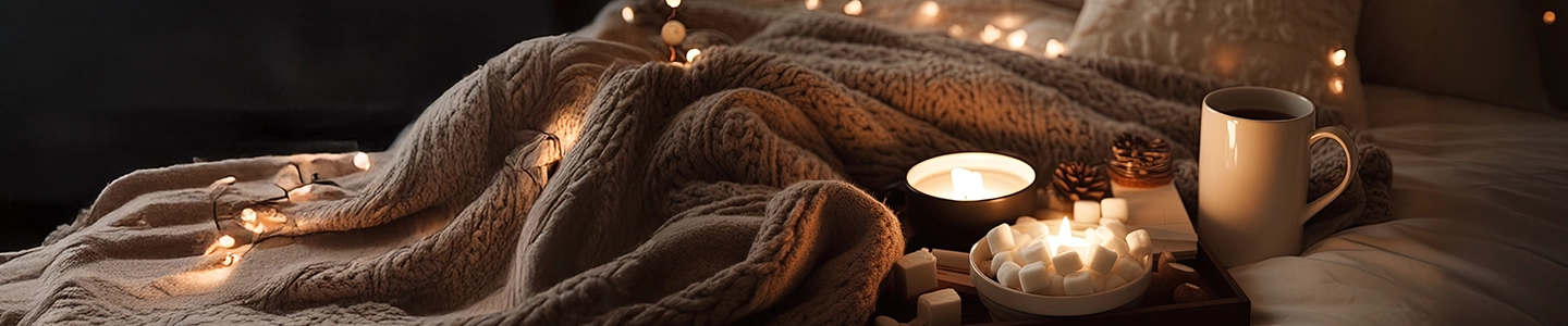 Gemütliche Szene mit einer warmen Decke, brennenden Kerzen, Lichterketten, einer Tasse und Marshmallows auf einem Bett, die eine entspannte Atmosphäre schaffen.